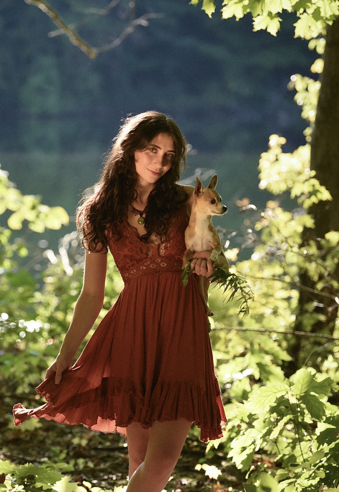 Young woman with long dark hair holding a small dog outdoors in a lush green setting, sunlight filtering through leaves.