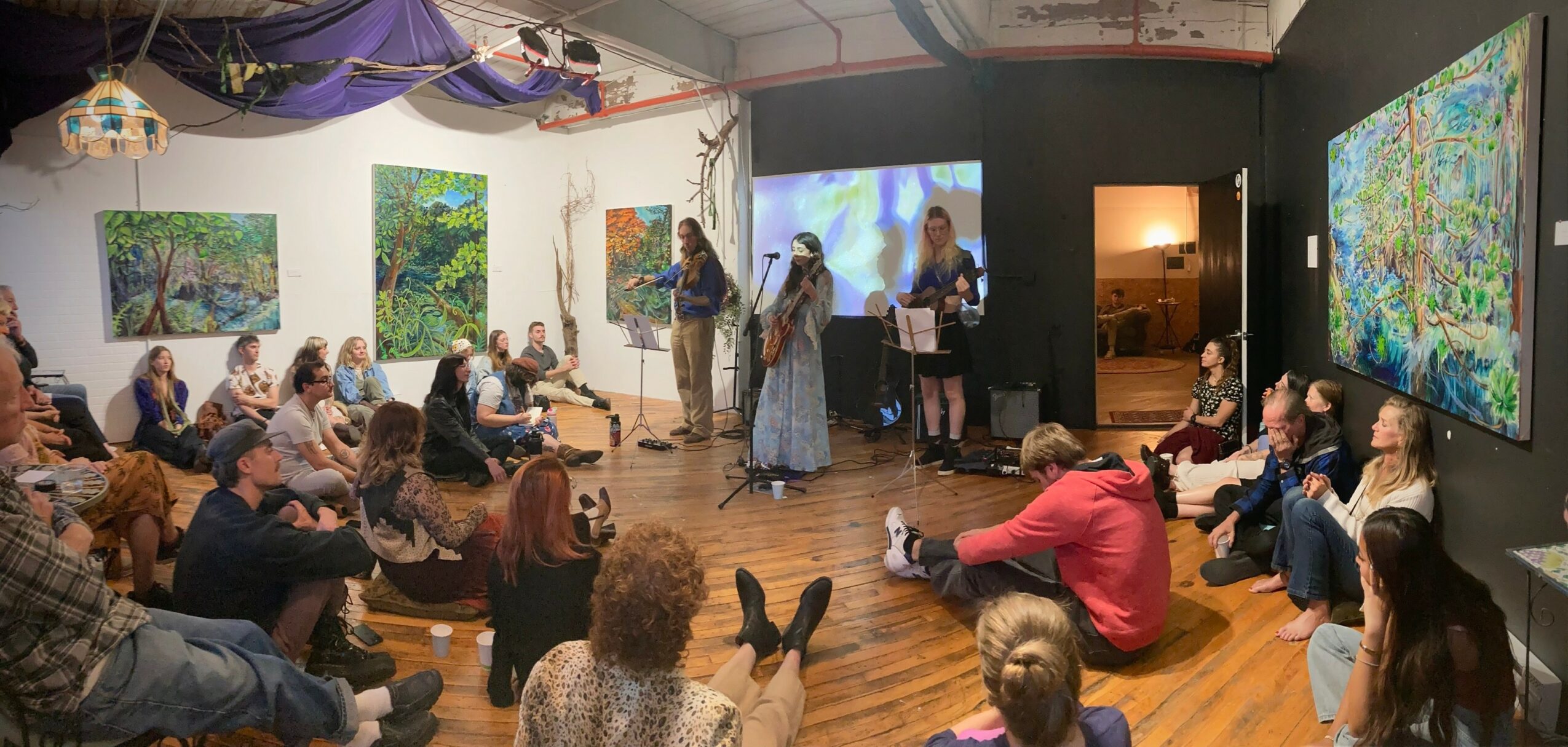 People seated in an art gallery watching a presentation with paintings on walls and a large screen, some taking photos.