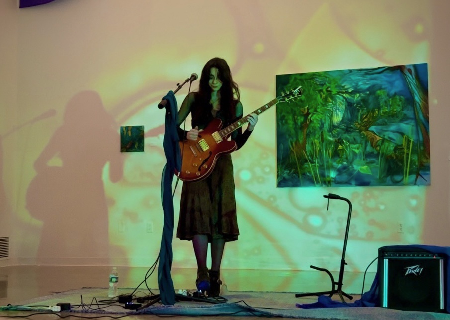 A woman with long dark hair plays an acoustic guitar on stage with colorful background lights.