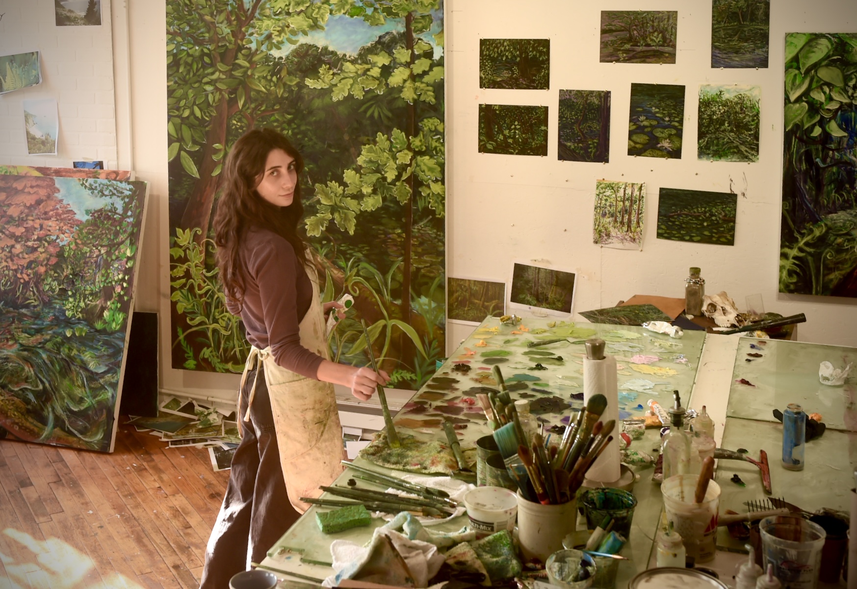 Woman in art studio with paintings of nature on walls and a table with art supplies.