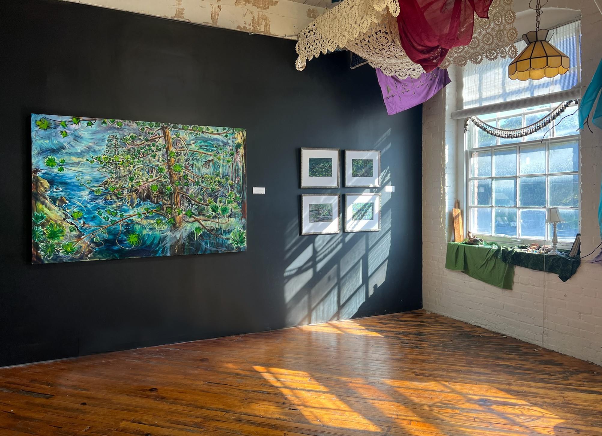Art gallery with paintings on dark wall, sunlight through window, wooden floor, and colorful fabric decorations hanging from ceiling.