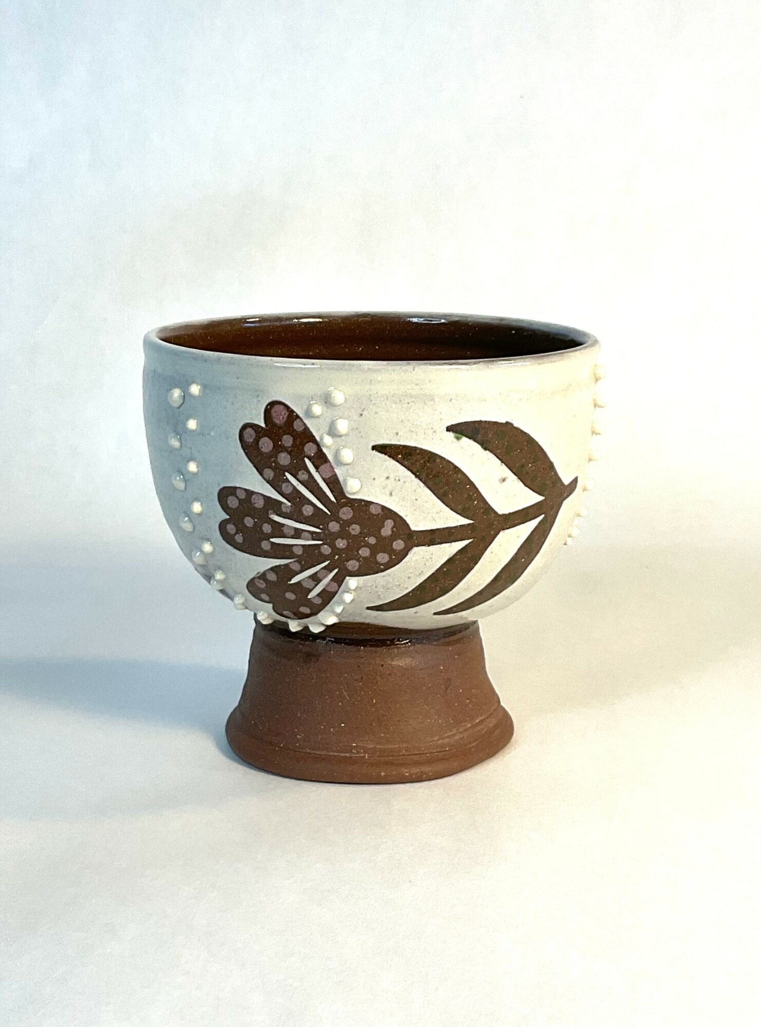 Decorative ceramic cup with a butterfly and leaf design, on a brown base, against a plain background.