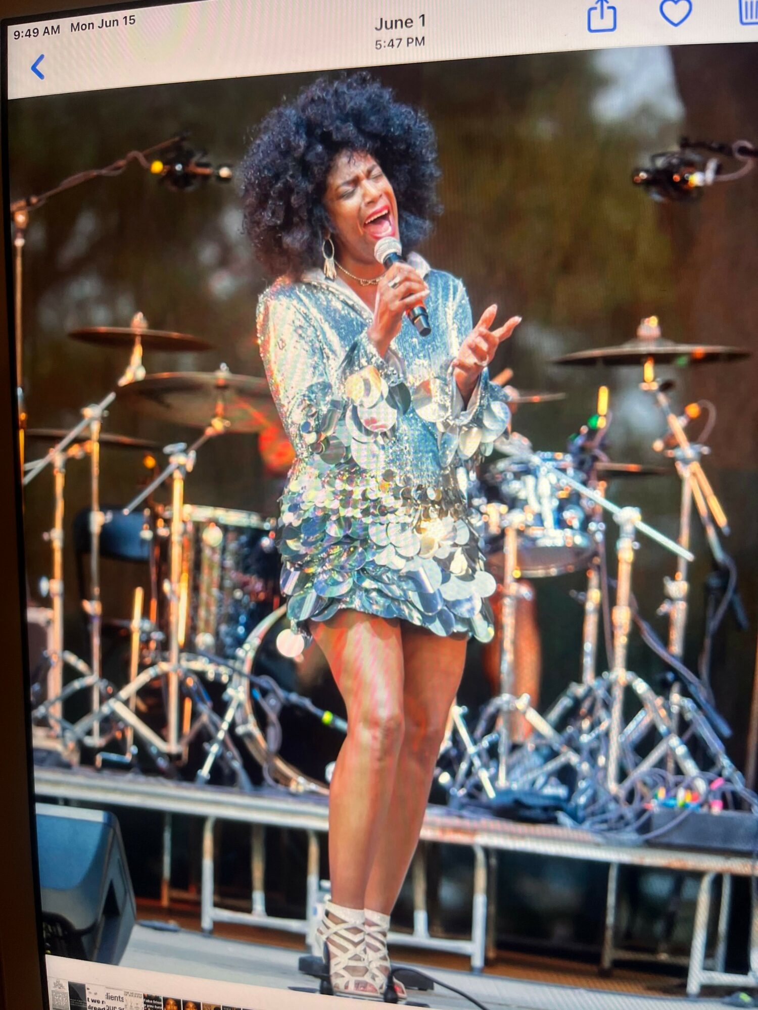 Performer on stage with a colorful outfit, holding a microphone, with stage equipment and lights in background.