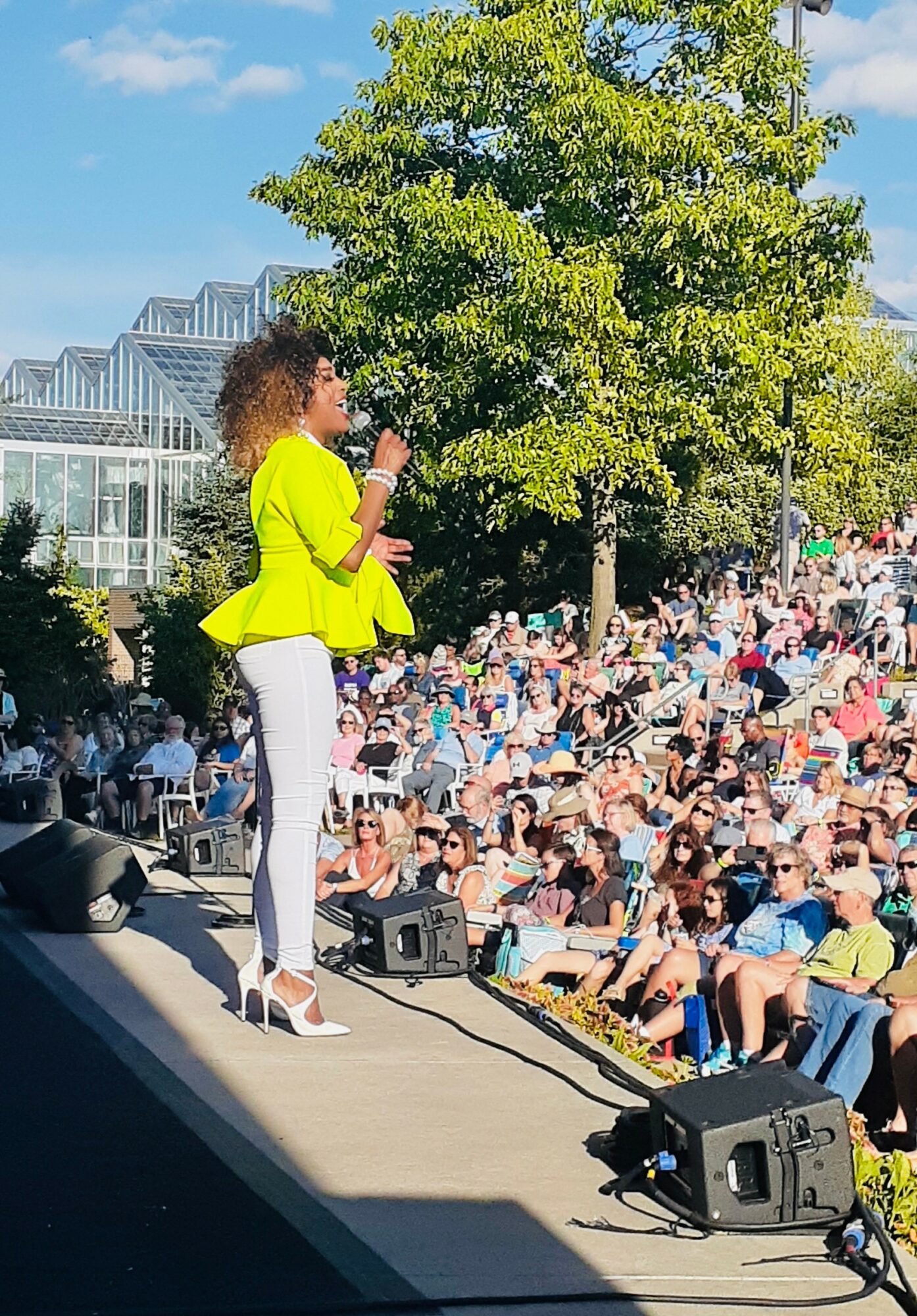 Woman in bright yellow jacket and white pants speaking to large outdoor crowd, green trees and blue sky in background.