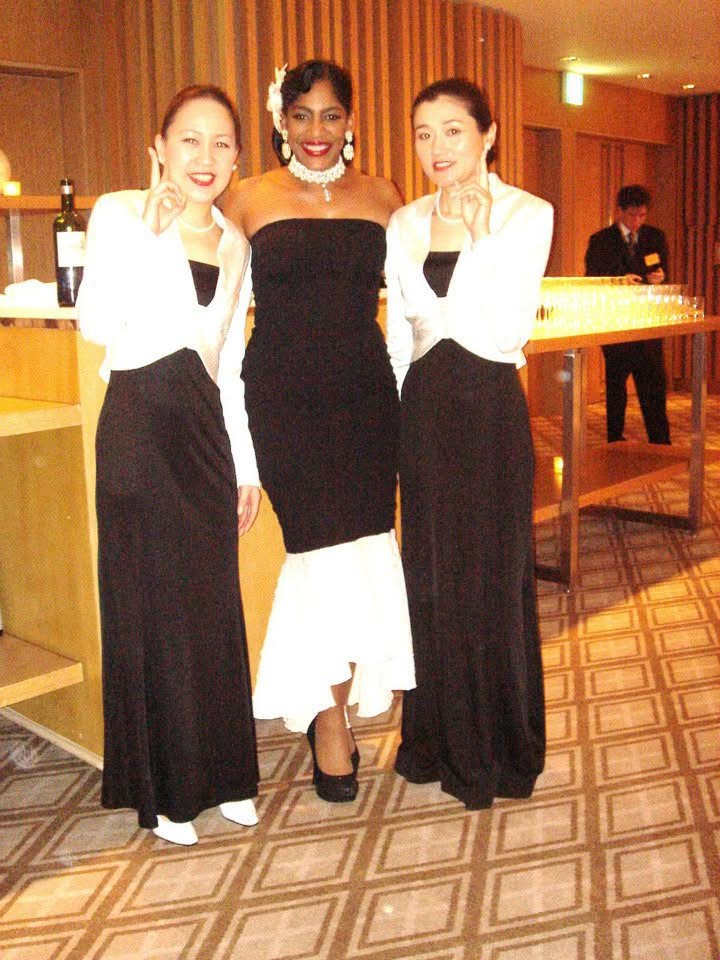 Three women in formal attire standing together in a room with warm lighting and wooden decor.