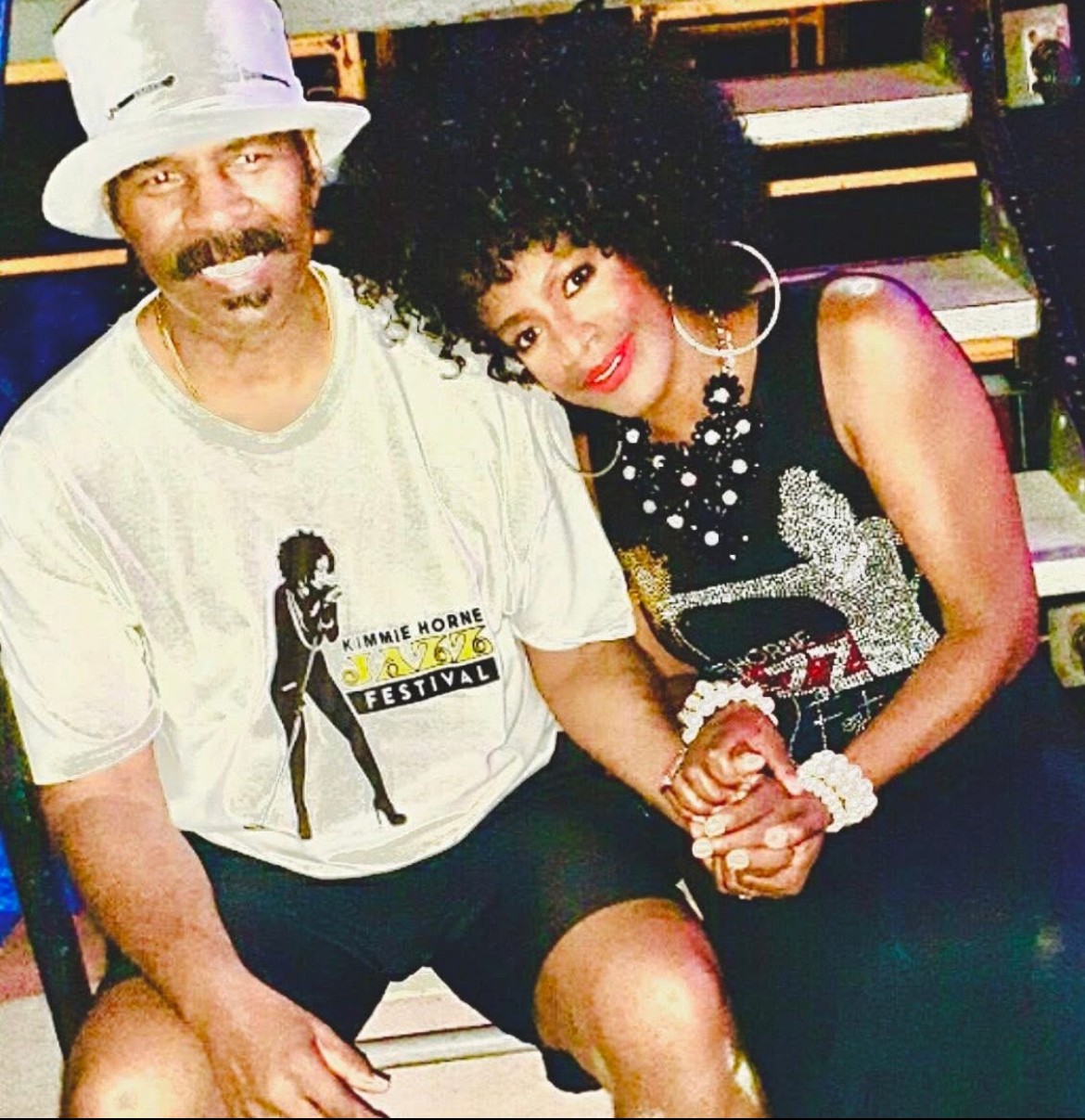 Two people sitting close together, holding hands, smiling, with a staircase in the background.