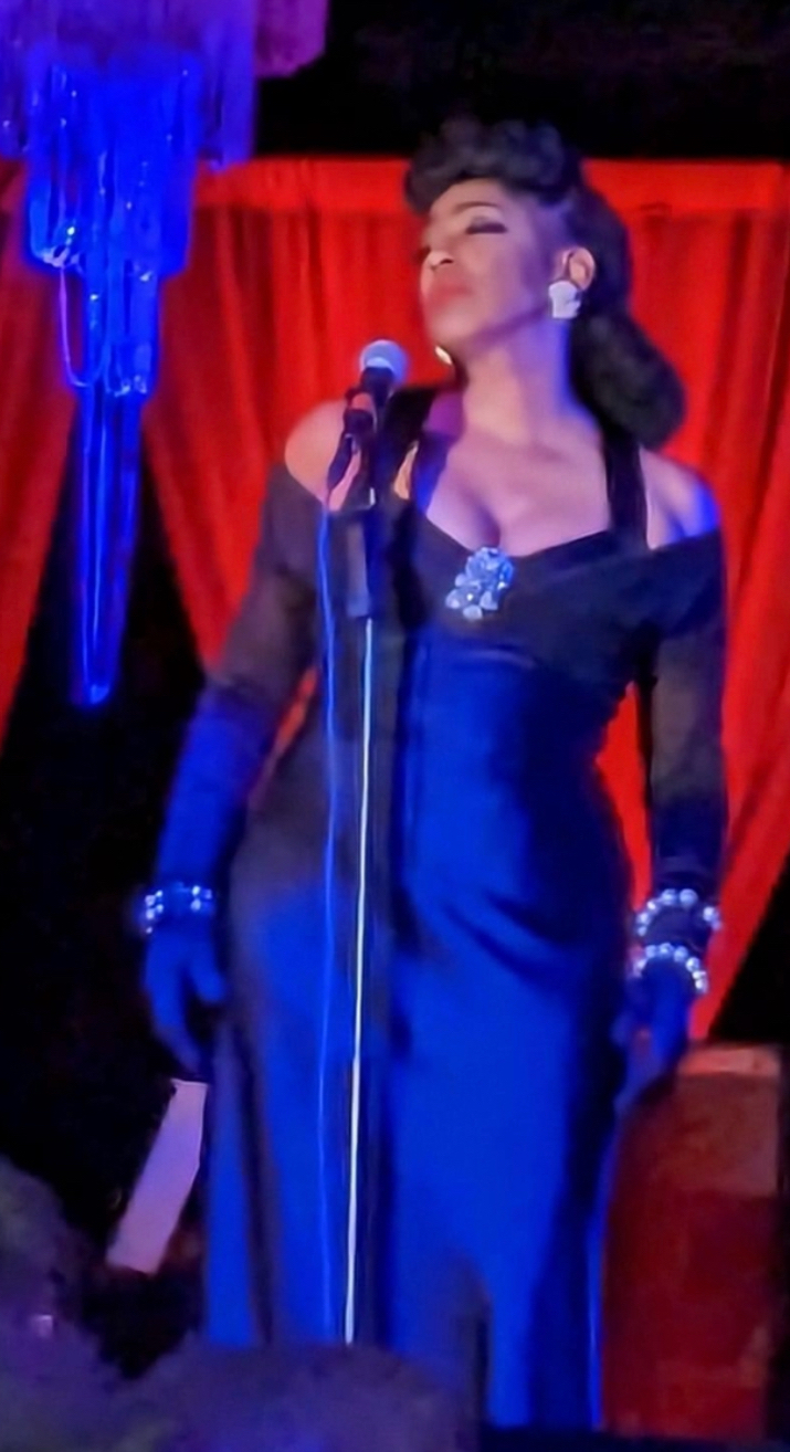 Woman singing into a microphone on stage with red curtains and blue decorations behind her.
