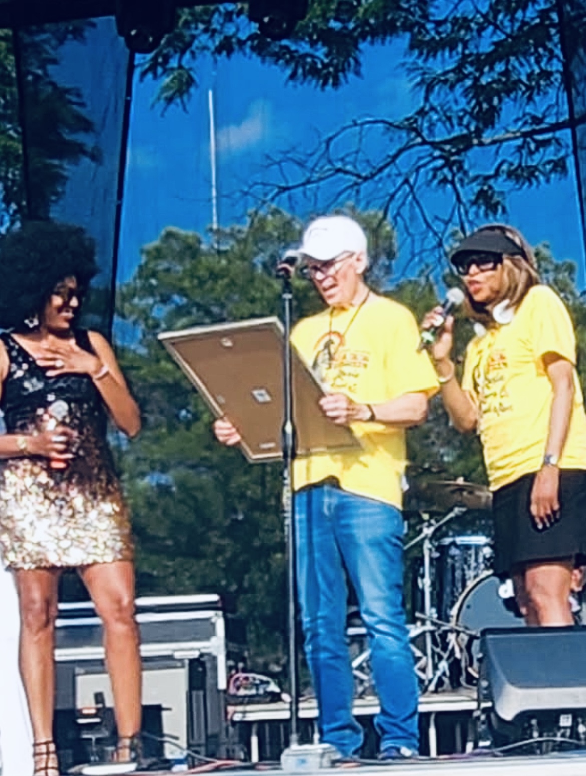 Three people on stage outdoors, two women and one man, with trees and equipment in the background, speaking into microphones.