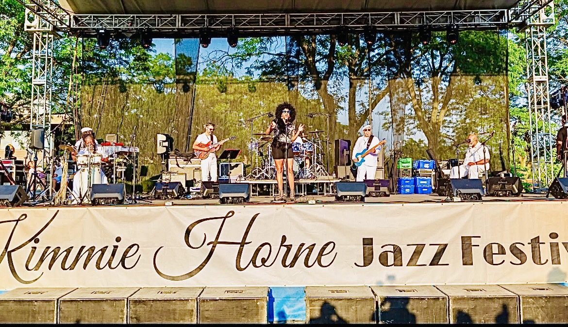 Musicians perform on stage at a jazz festival outdoors with trees in background.
