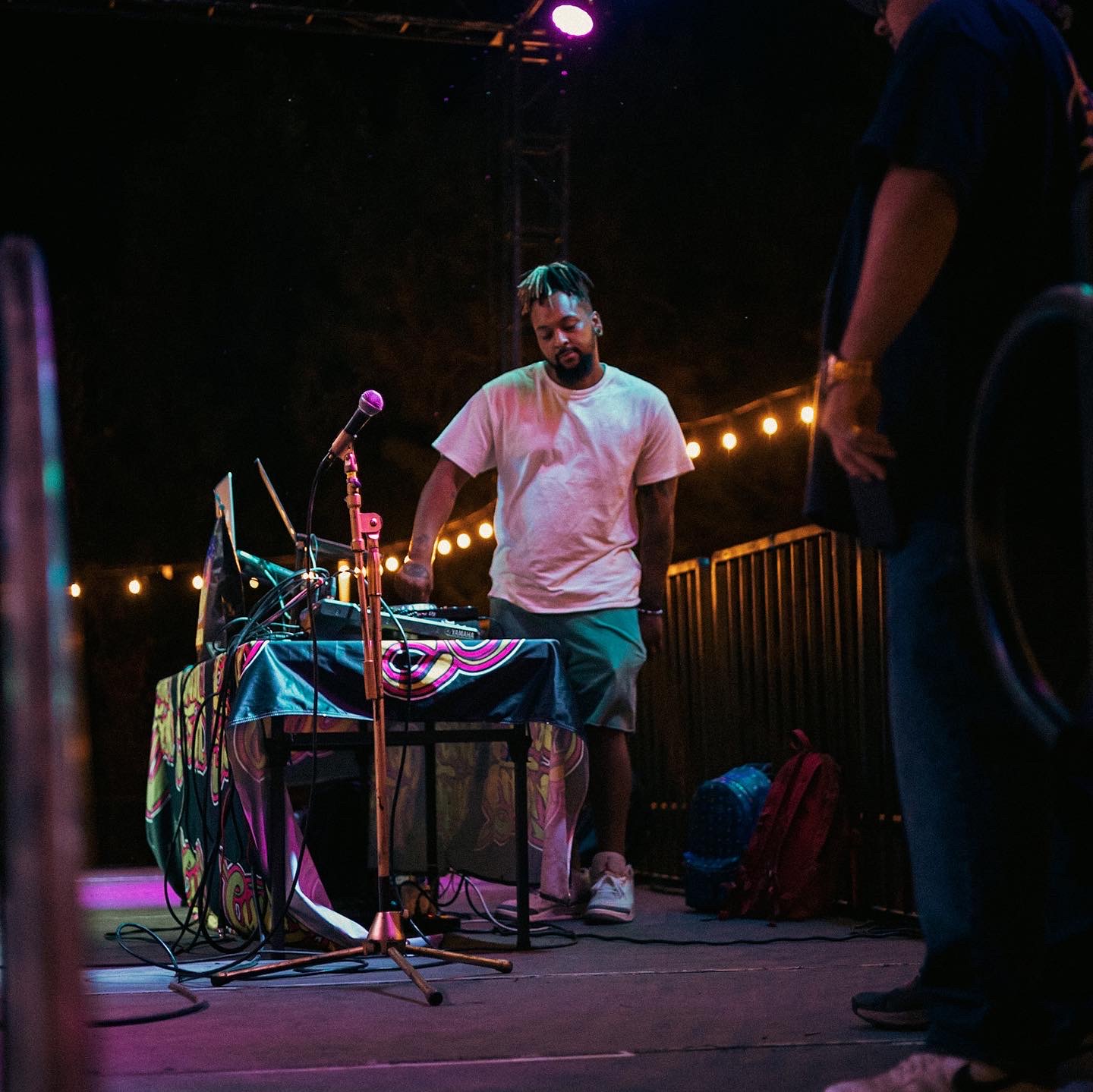 Man with beard and white t-shirt standing behind a table with electronic equipment on stage, with string lights in background.