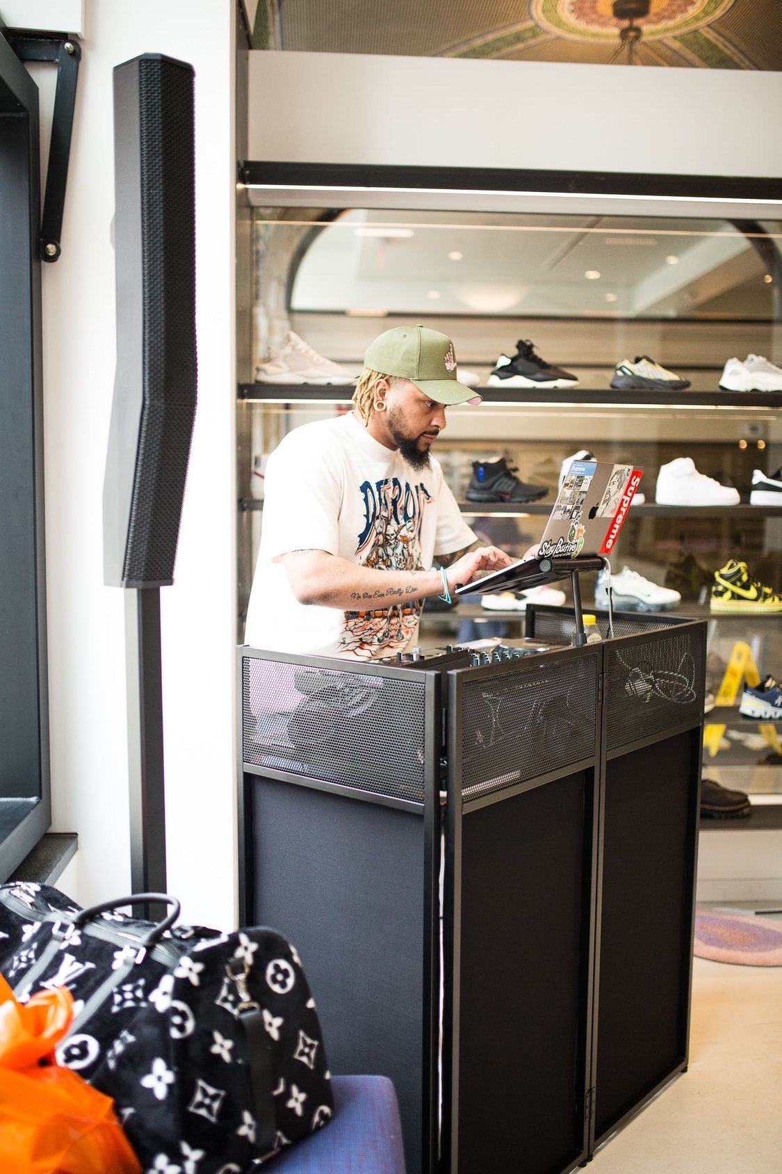 A man with a green cap and white shirt stands behind a checkout counter in a shoe store, browsing items on a screen.
