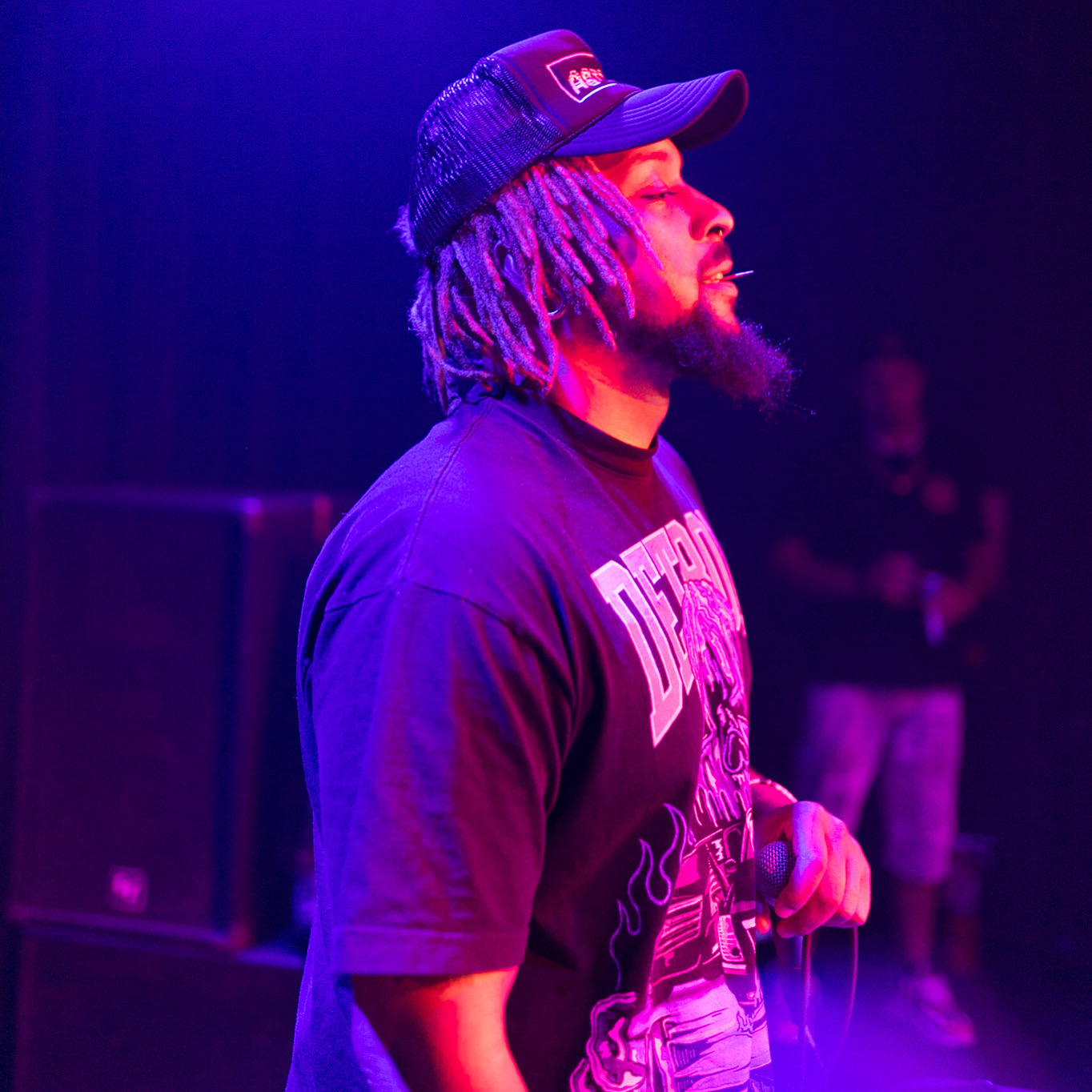 Side view of a man with dreadlocks, wearing a cap and a graphic T-shirt, illuminated by purple and pink stage lights.
