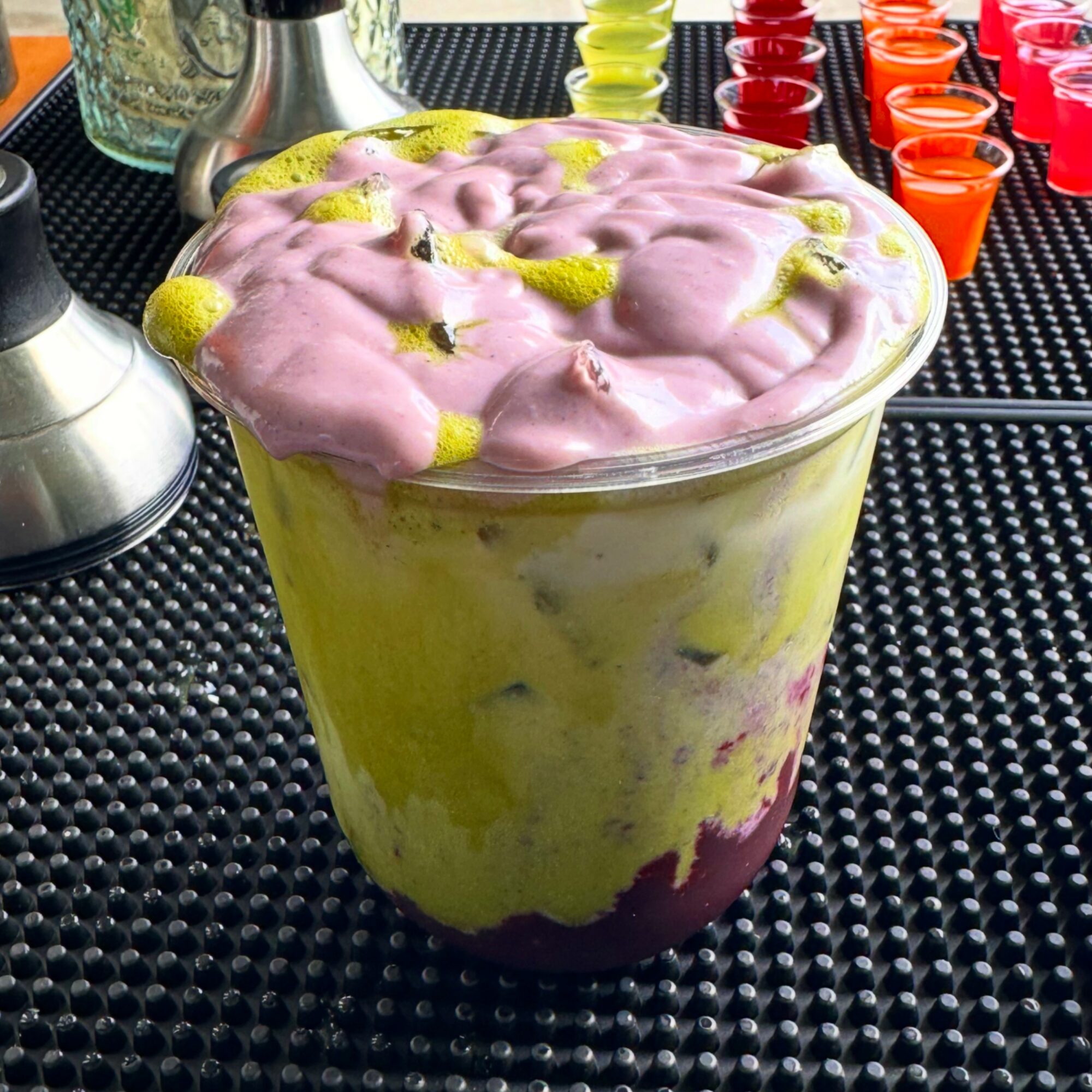 A large glass container filled with layered yellow, pink, and purple dessert topped with pink foam, on a black textured surface.