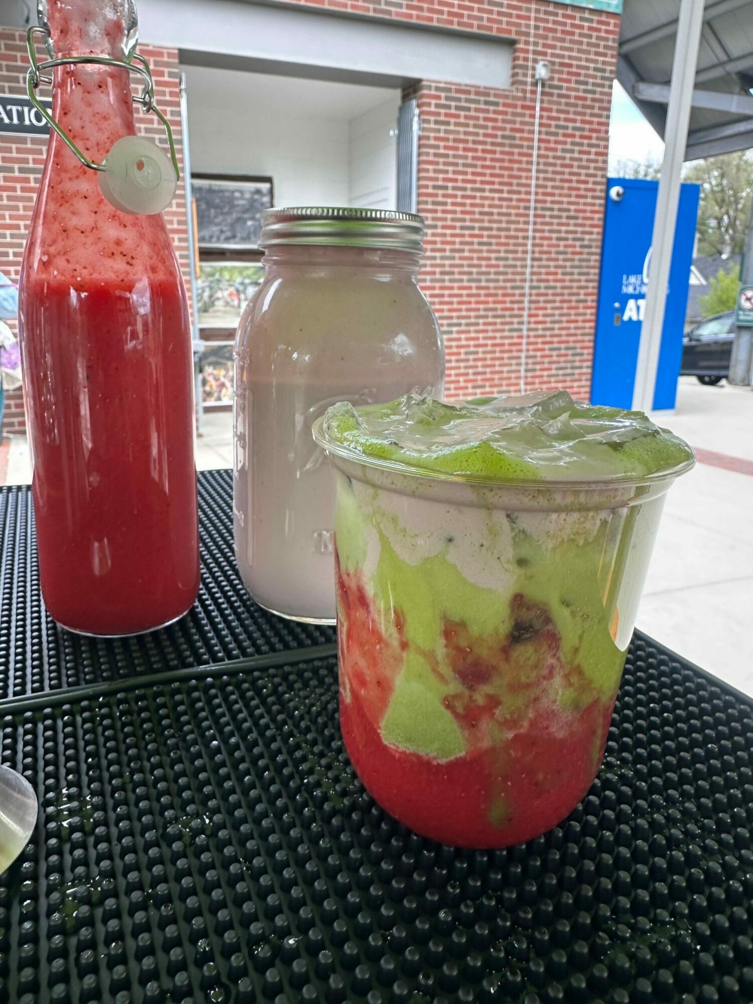 Two mason jars with colorful smoothies on a black surface outdoors, one with a straw and a brick building in the background.
