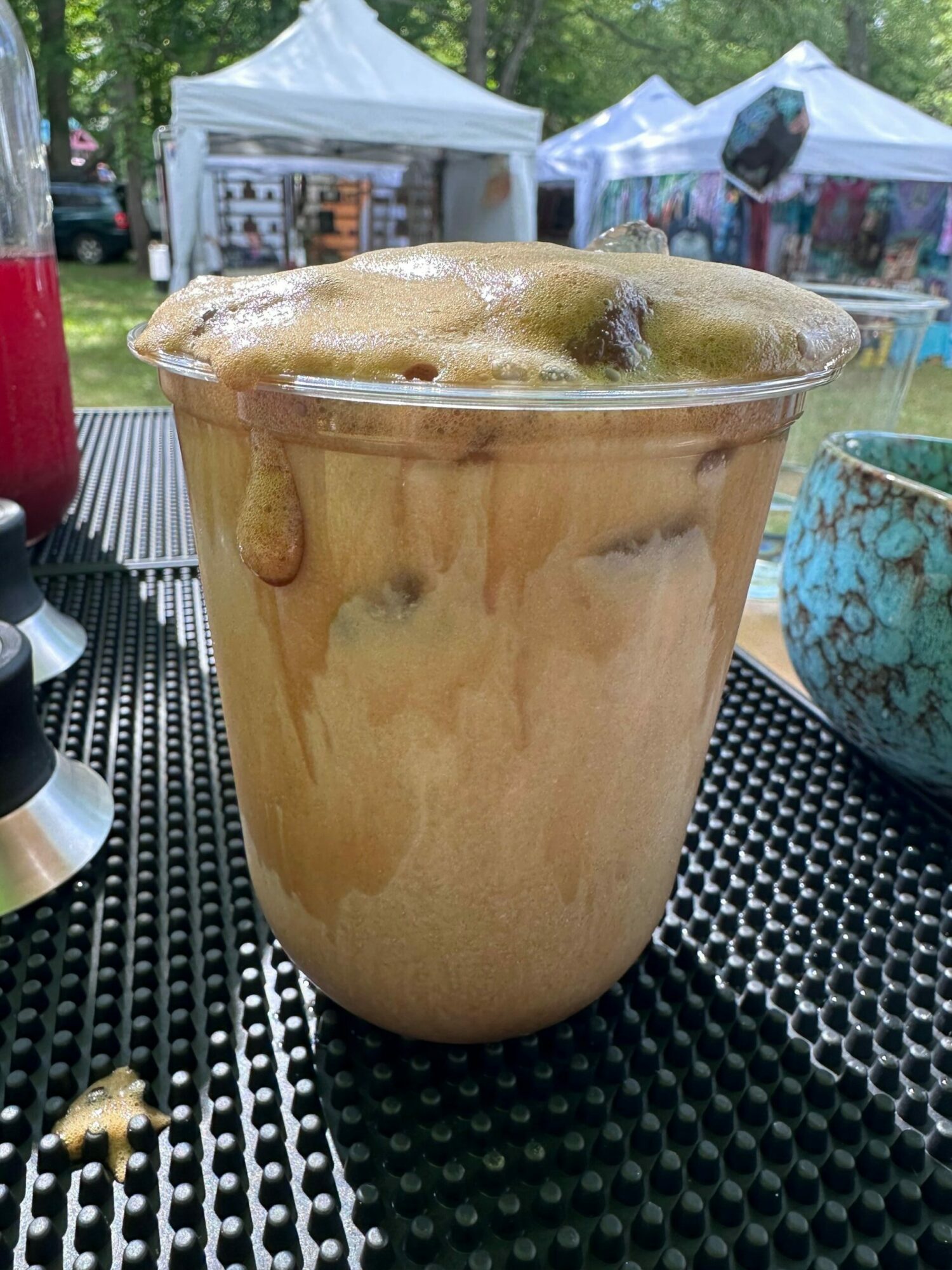 A glass of iced coffee with foam on top, placed on a black textured surface outdoors.