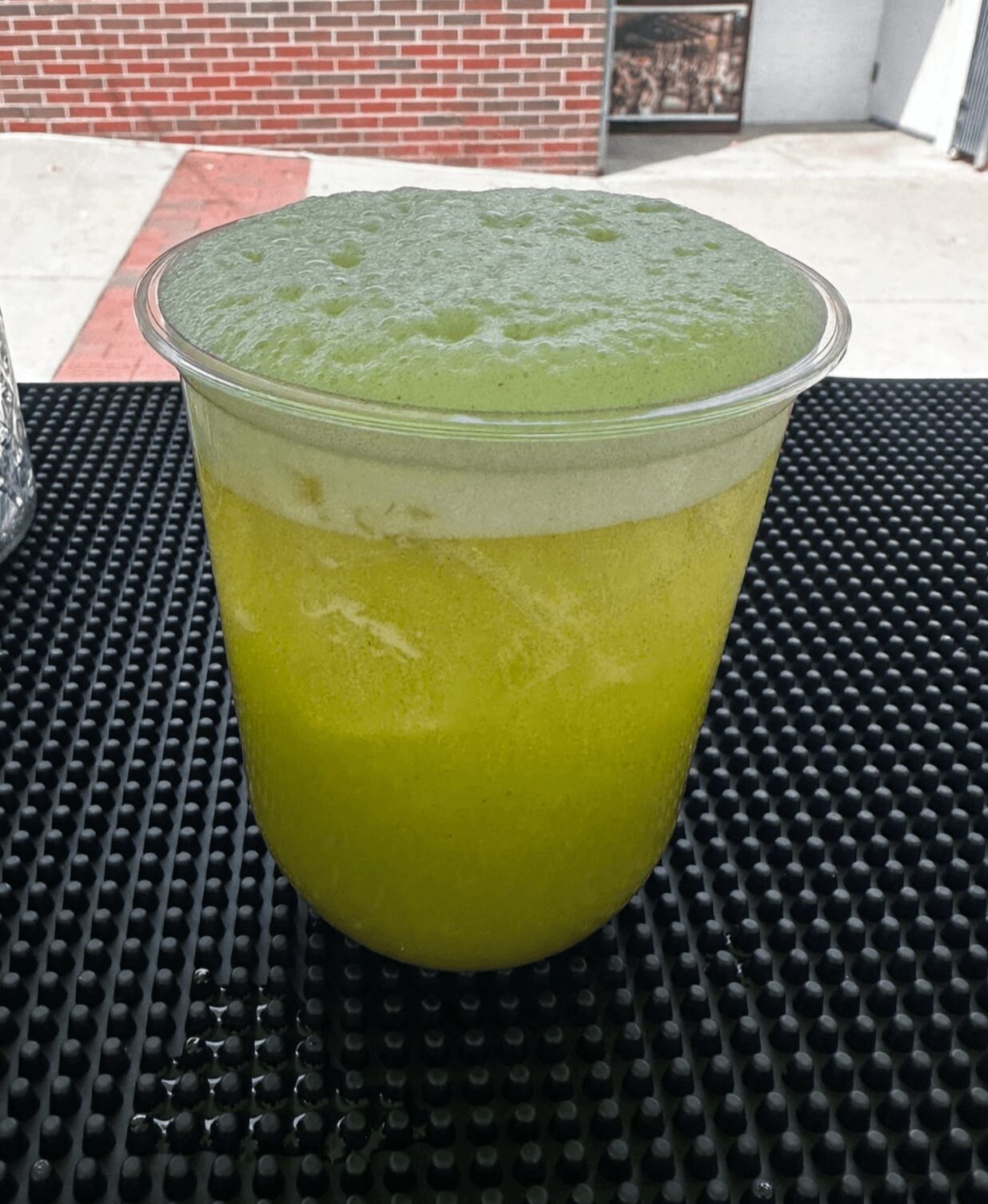 Clear plastic cup filled with bright green slushy beverage on black textured surface, outdoor background with brick wall and sidewalk.