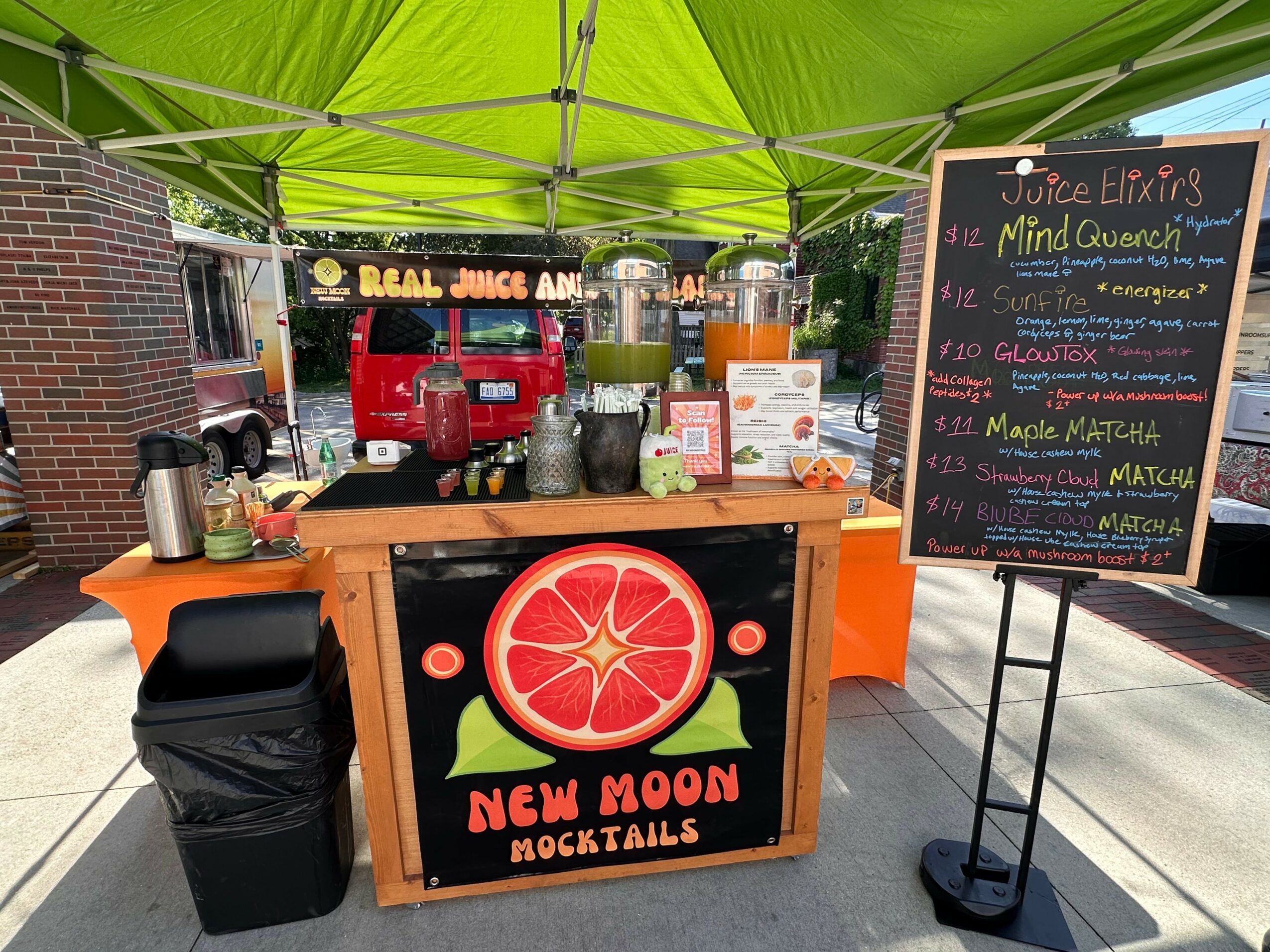 Juice stand with a green canopy, colorful drinks, a chalkboard menu, and a sign with a red citrus fruit and 'New Moon Cocktails'.