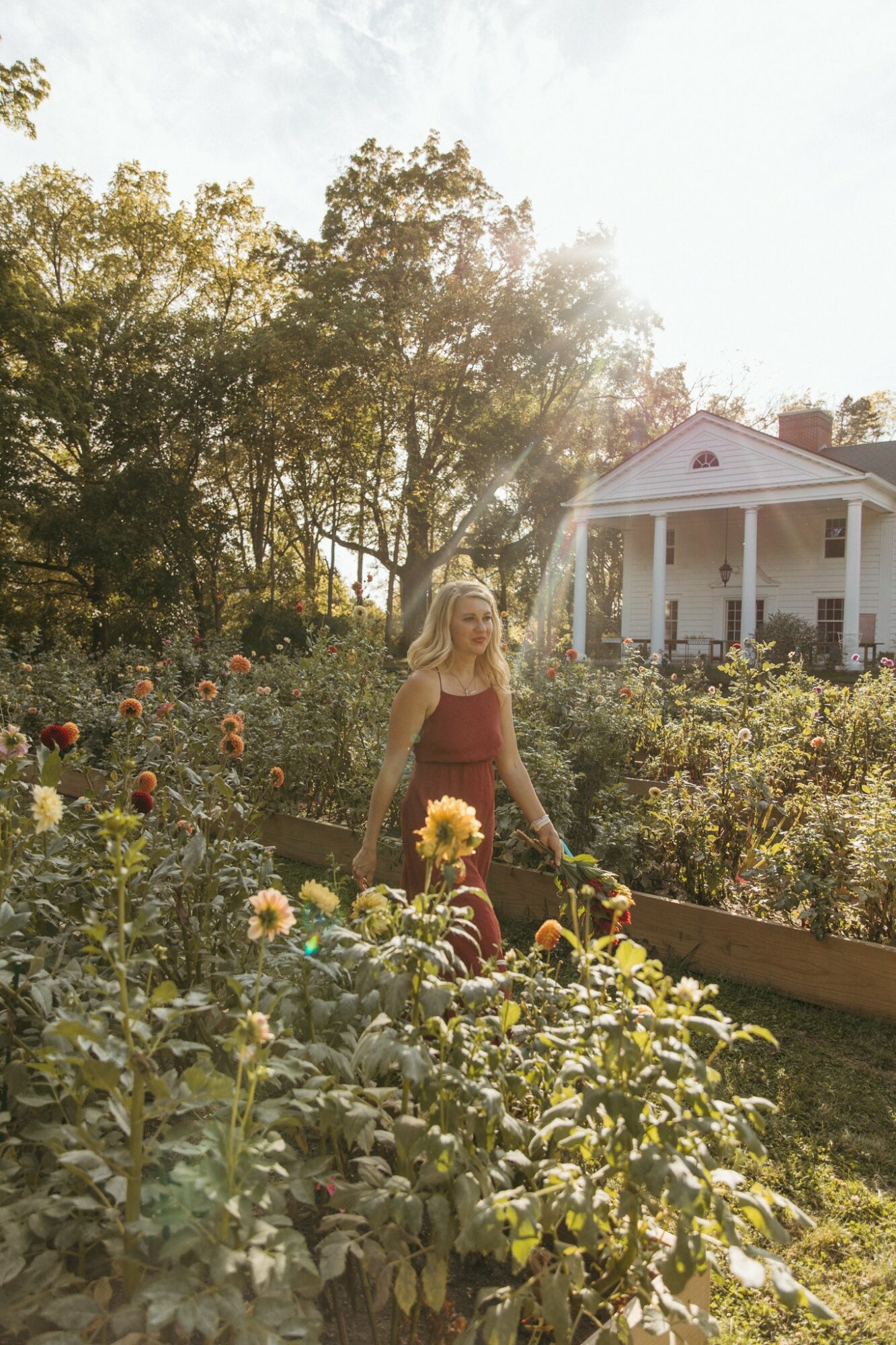 Woman with long blonde hair in a garden with colorful flowers, sunlight shining, and a white house in the background.