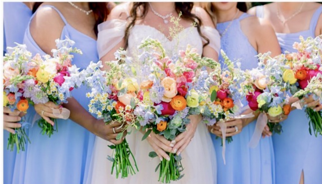 Group of women in blue dresses holding colorful flower bouquets, with a woman in the center wearing a white dress.
