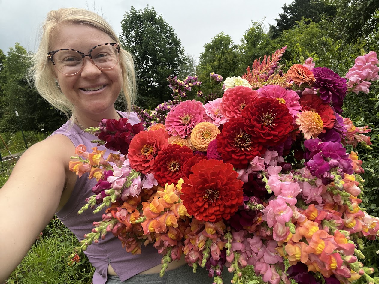 Smiling woman with glasses holding a large colorful flower bouquet outdoors, trees and sky in background.