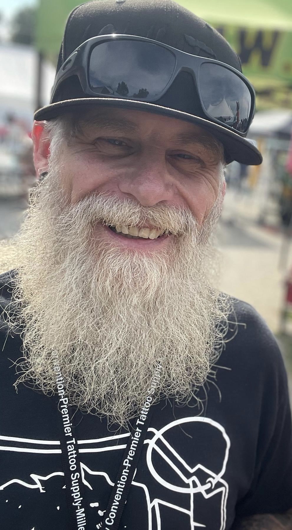 Smiling man with a long beard wearing a black cap and sunglasses outdoors.