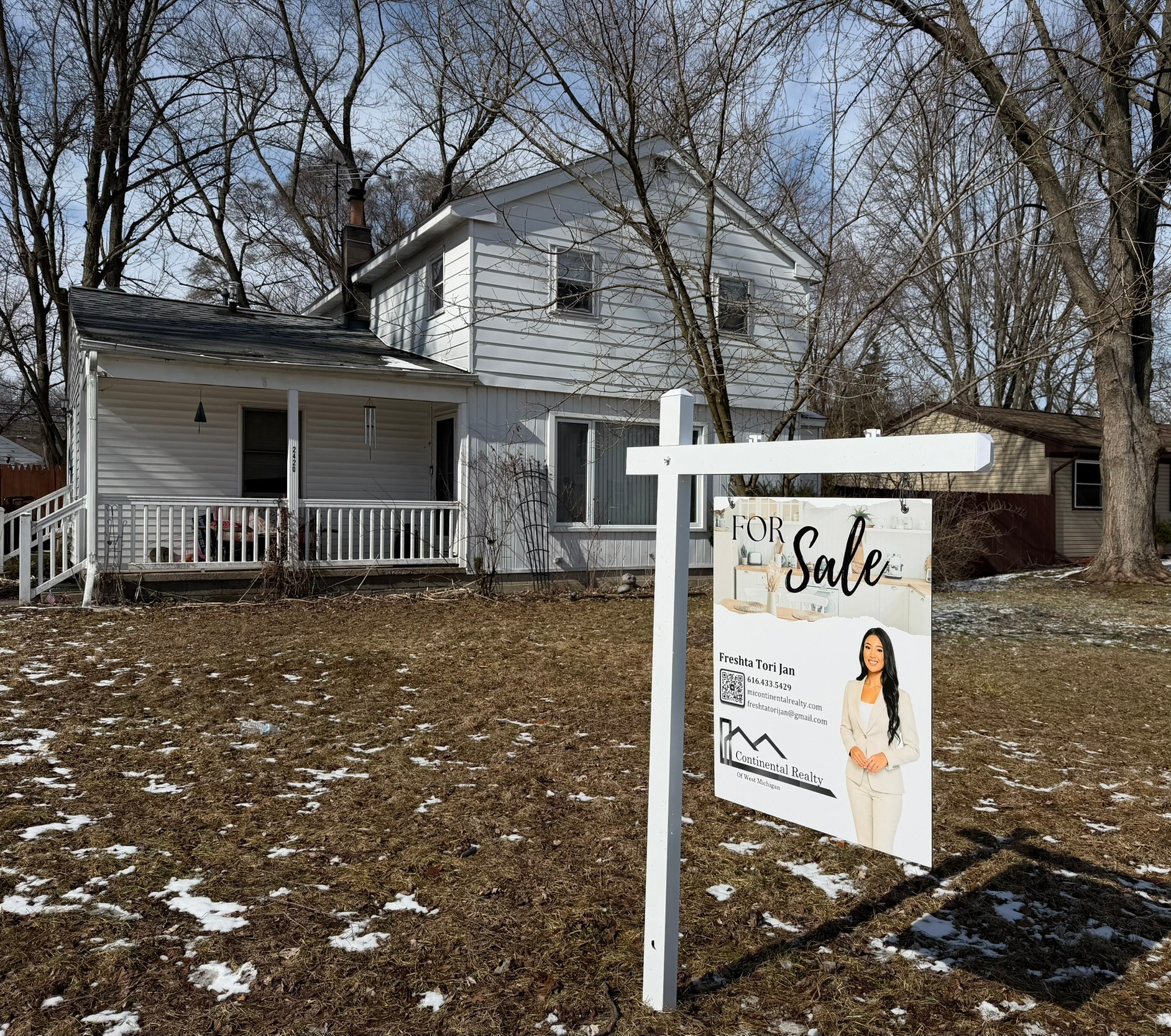 House with a porch and a for sale sign in front, trees in the background, some snow on the ground.