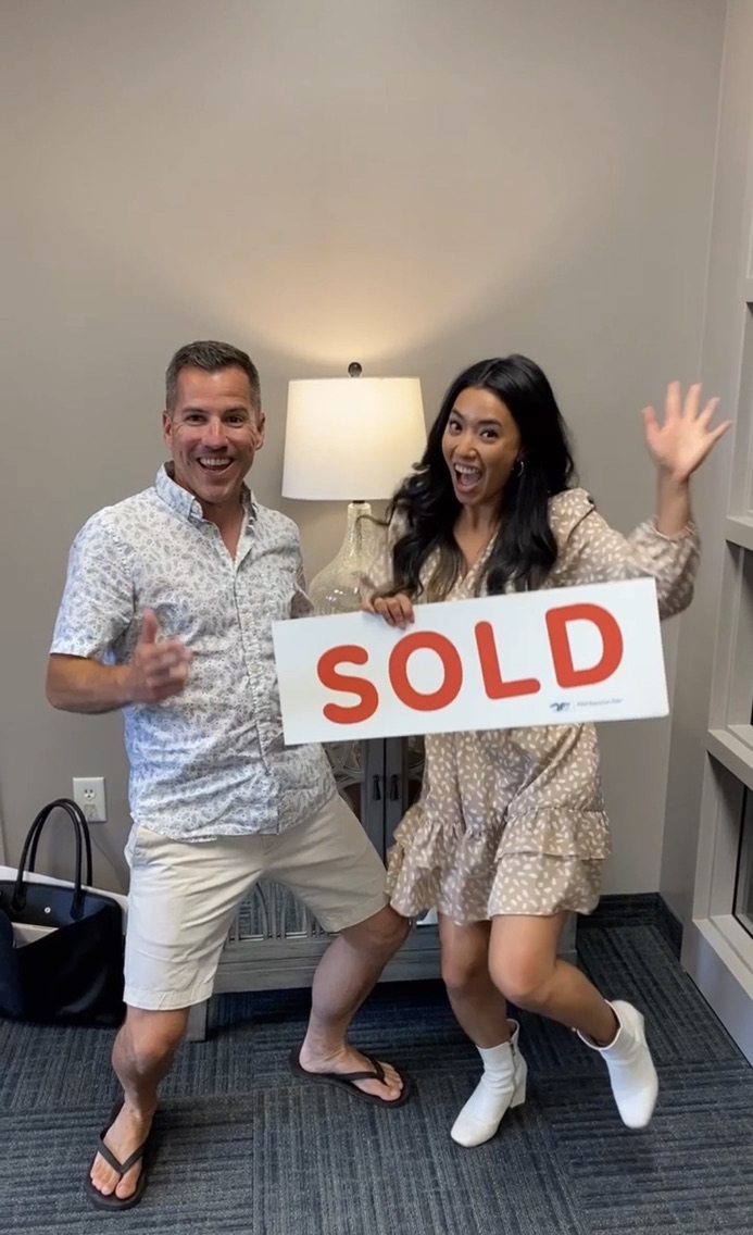 Two people smiling, one holding a 'SOLD' sign, in a room with a lamp and bookshelf, celebrating a sale.