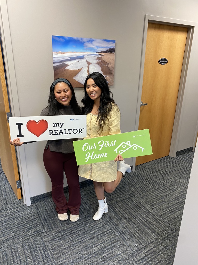 Two women standing indoors, holding signs, smiling, with a door and a landscape photo in the background.