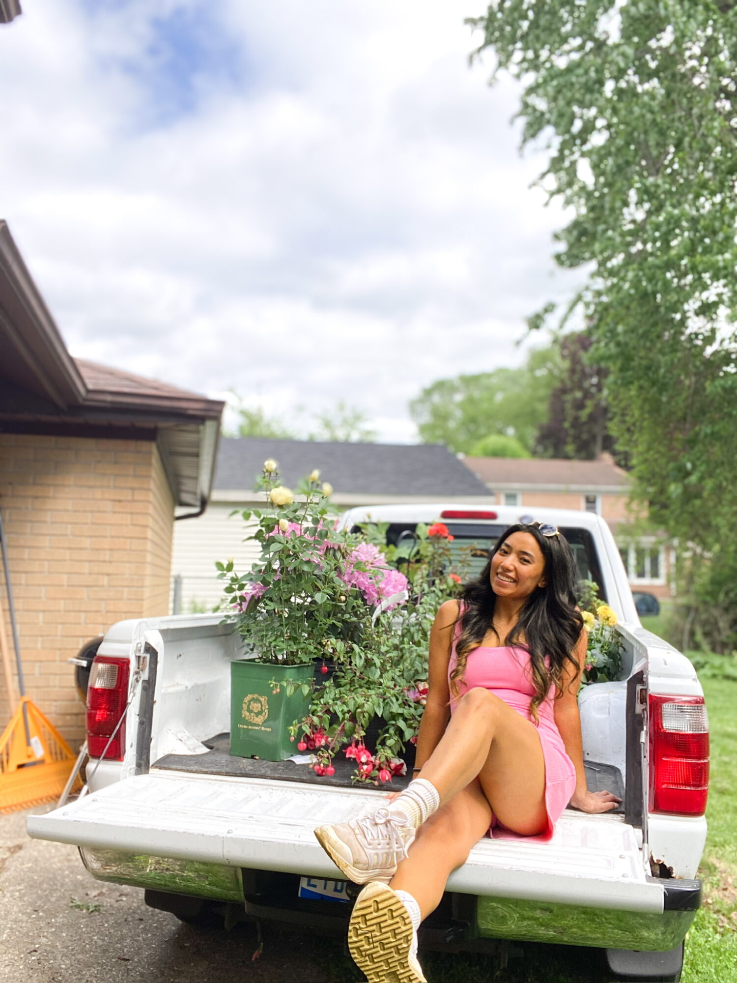 Young woman in pink dress sitting on the back of a white pickup truck, smiling outdoors with trees and buildings nearby.