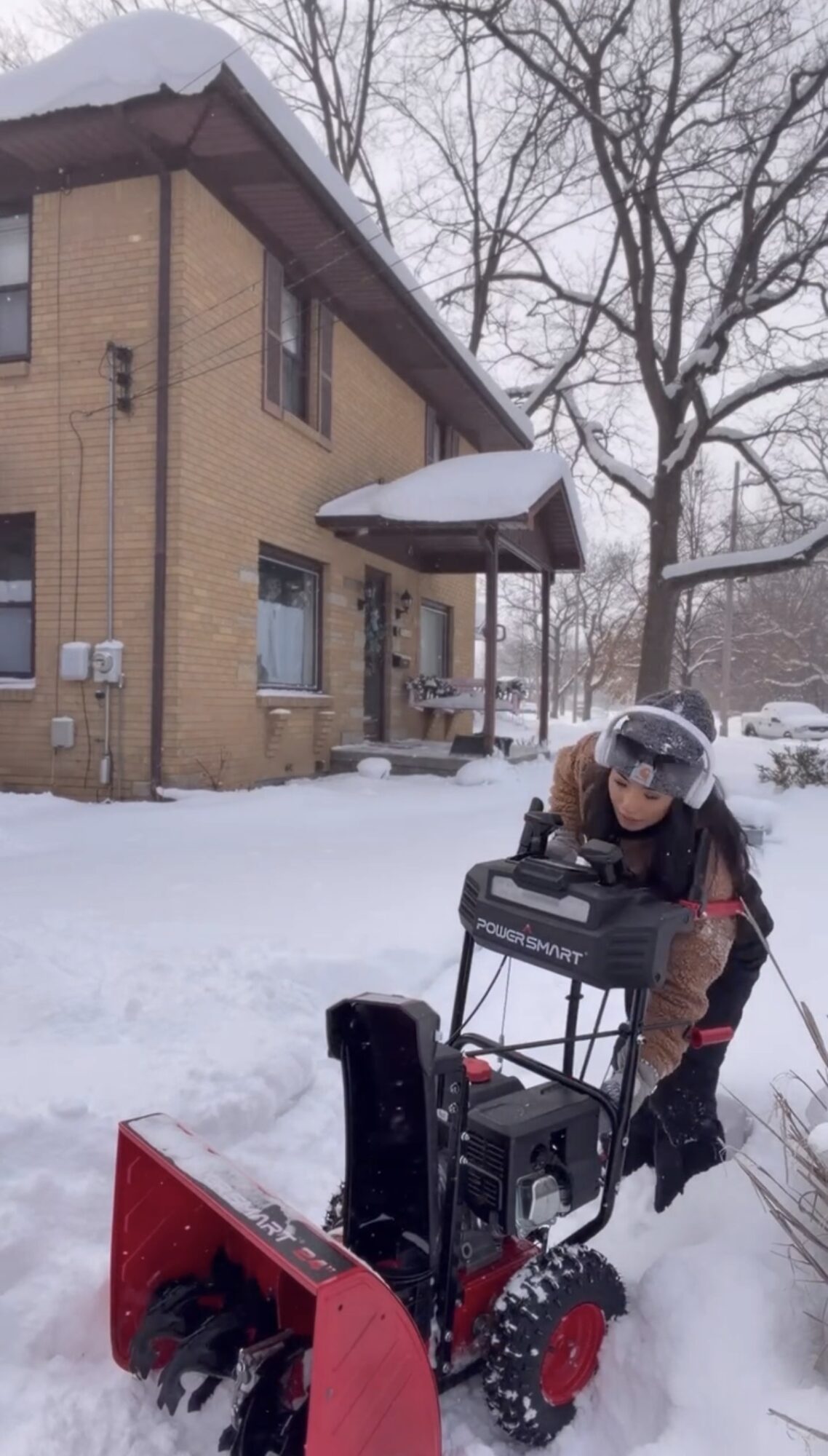 Person operating a snowblower outside a brick house in snowy weather, with trees in the background.