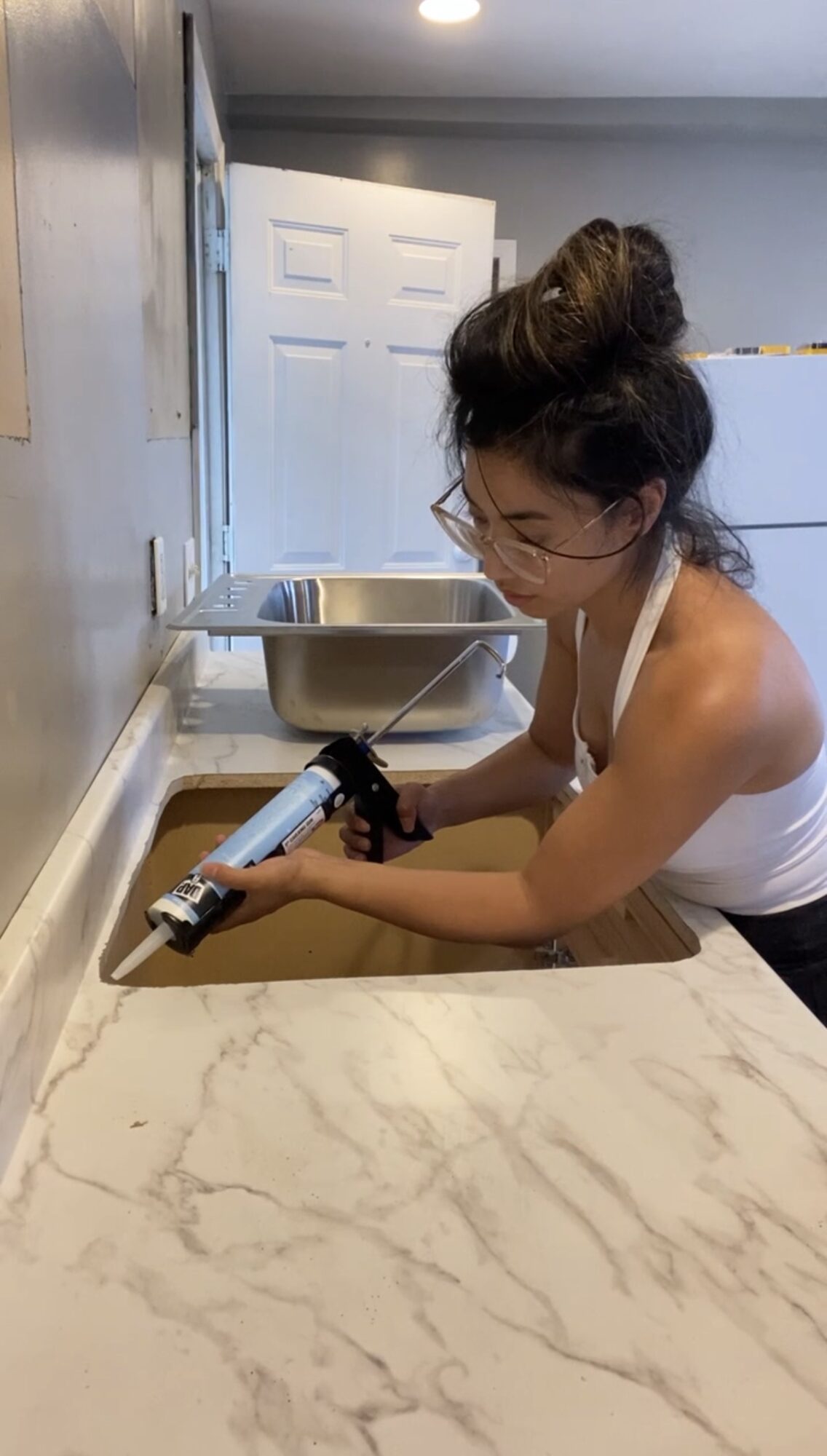 Person with glasses and a bun using a caulking gun on a countertop, in a kitchen with a sink and door behind.