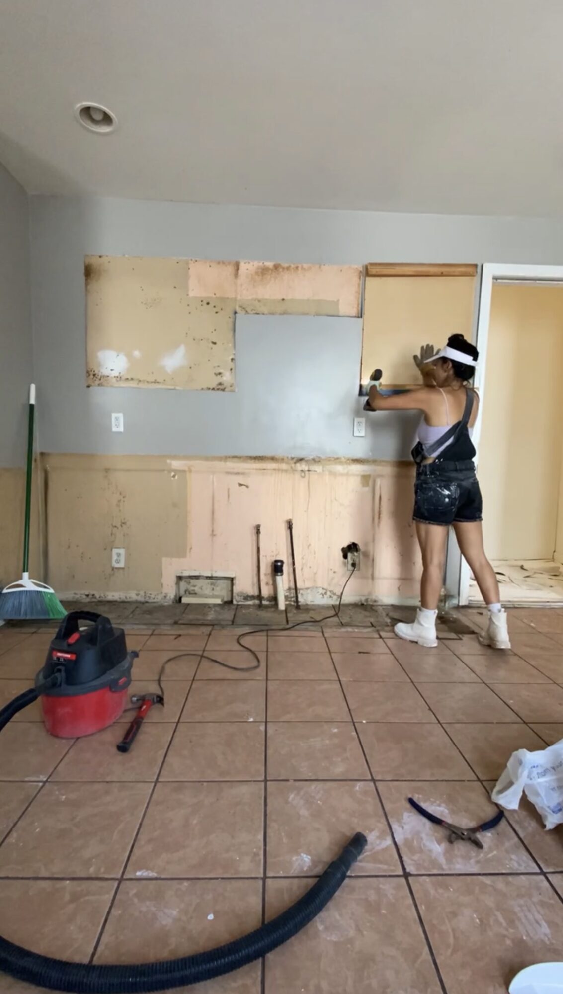 Person working on kitchen wall with tools, partially removed cabinets, and cleaning supplies on tiled floor.
