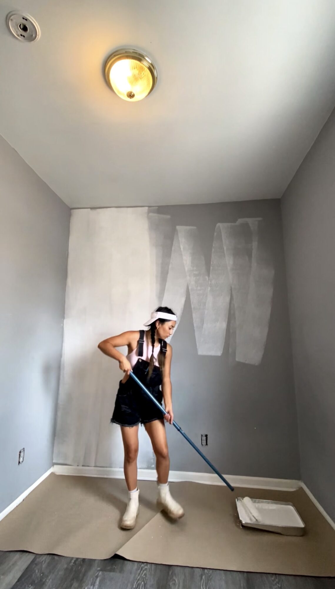 Person painting wall with roller in a room, ceiling light on, partially painted wall, floor covered with protective sheet.