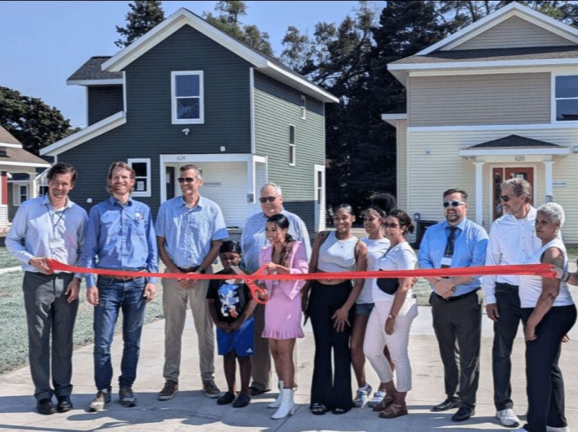 Group of people at a ribbon-cutting event in front of new houses, holding a red ribbon, smiling, sunny day.