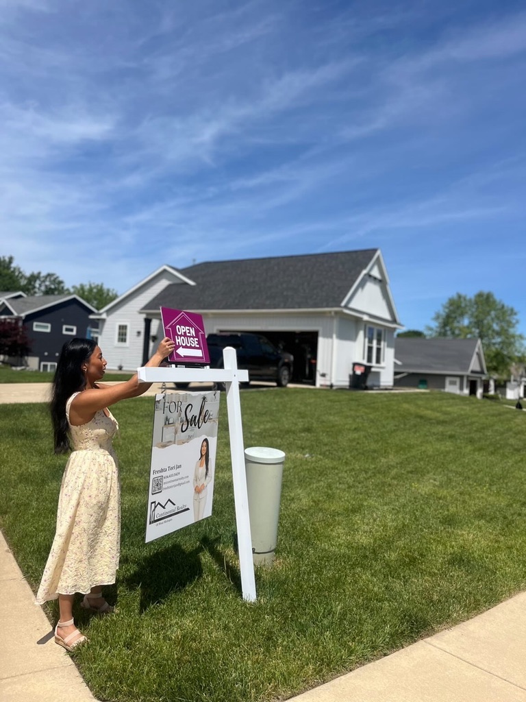 Woman placing a purple sign in front of a house with a garage, on a grassy lawn under a blue sky.
