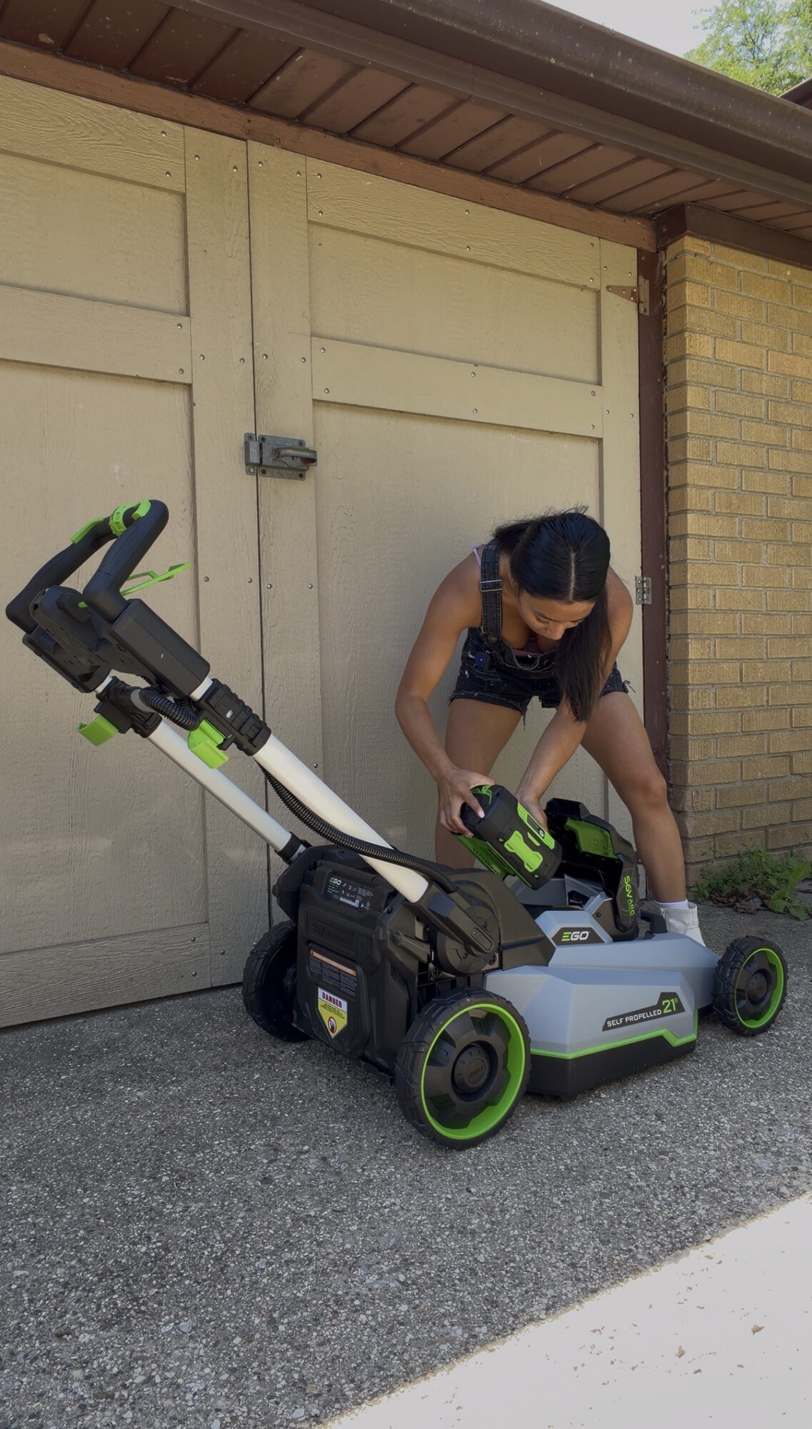Person adjusting a robotic lawn mower outside a garage door, with a brick wall nearby.