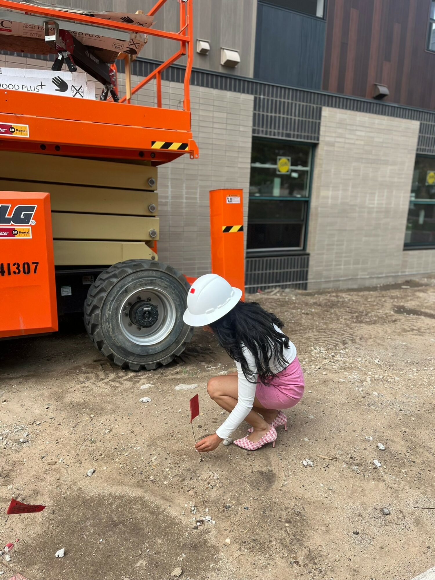 Child wearing a white hat and pink pants crouching on dirt ground, holding a small red flag near construction equipment outside building.