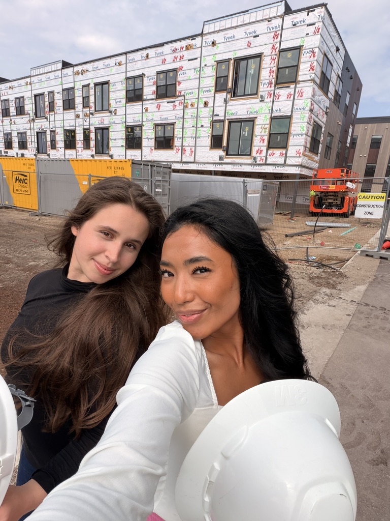 Two women taking a selfie outdoors in front of a building under construction, with construction equipment and barriers visible.