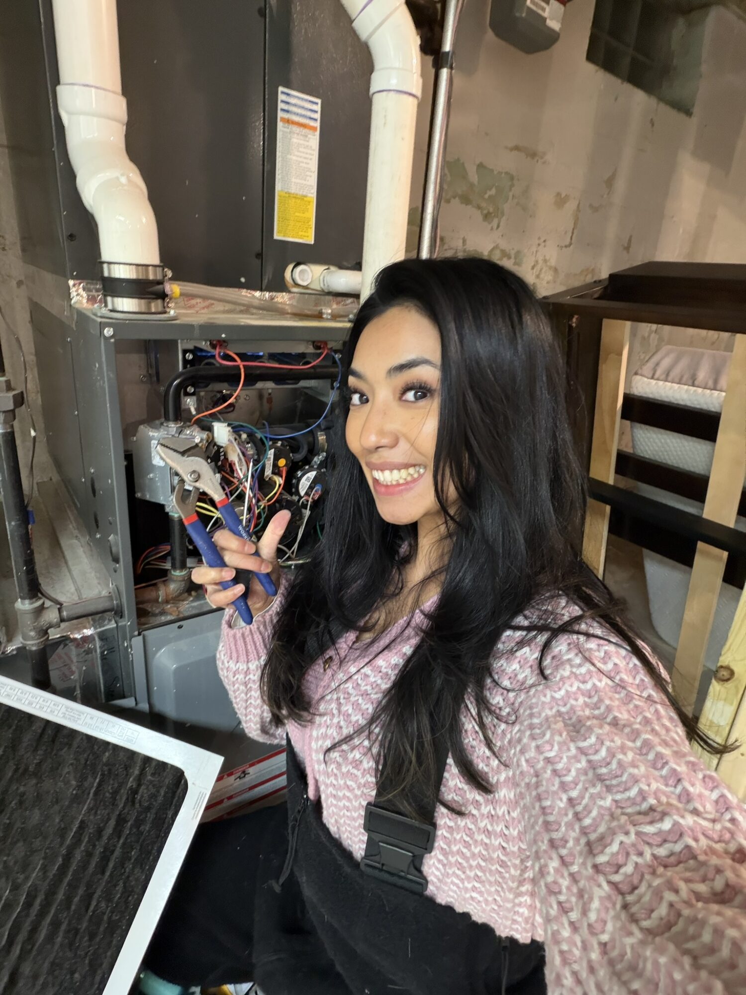 Young woman with long dark hair smiling, holding tools near an open mechanical or electrical panel, in a room with pipes and furniture.
