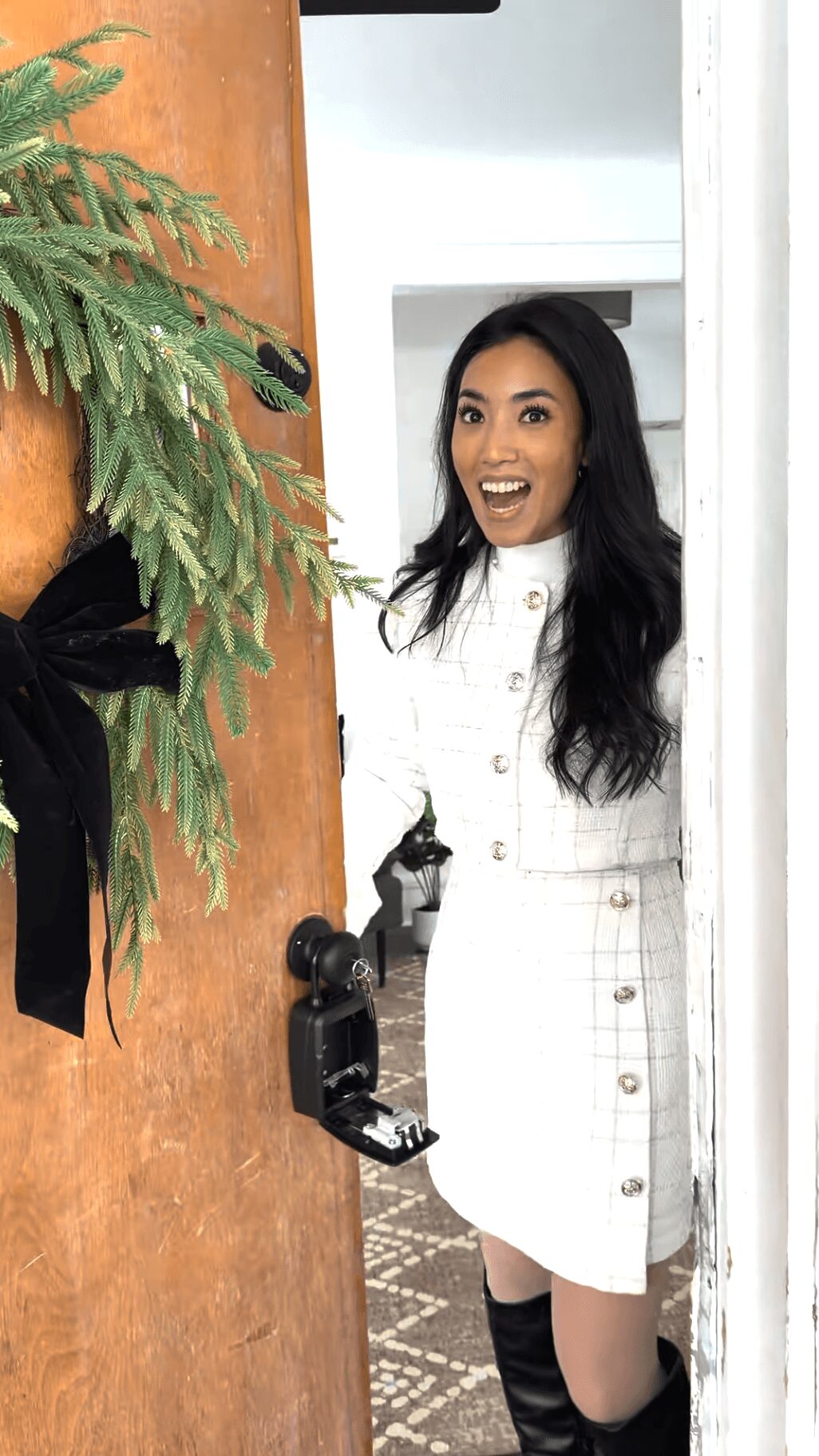 Woman with long dark hair smiling, wearing a white dress, standing indoors near a white wall.
