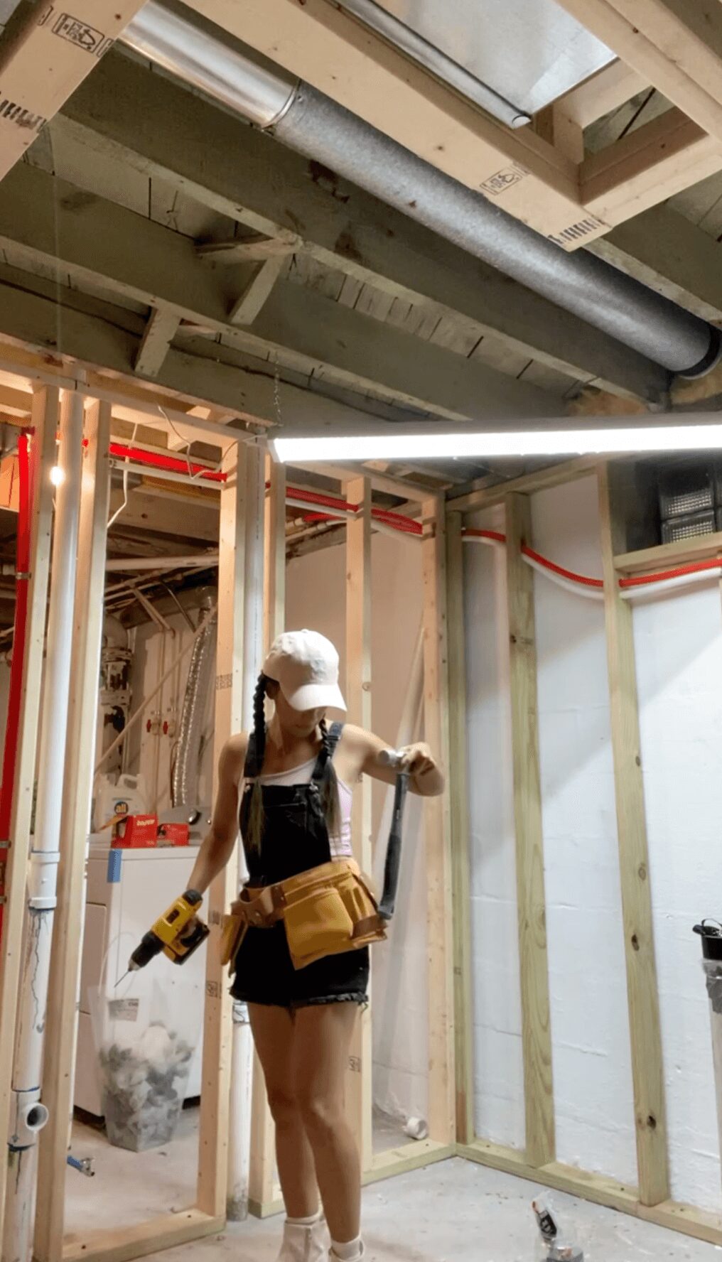Person wearing a white cap, black top, and shorts, working in a construction site with wooden framing and pipes.