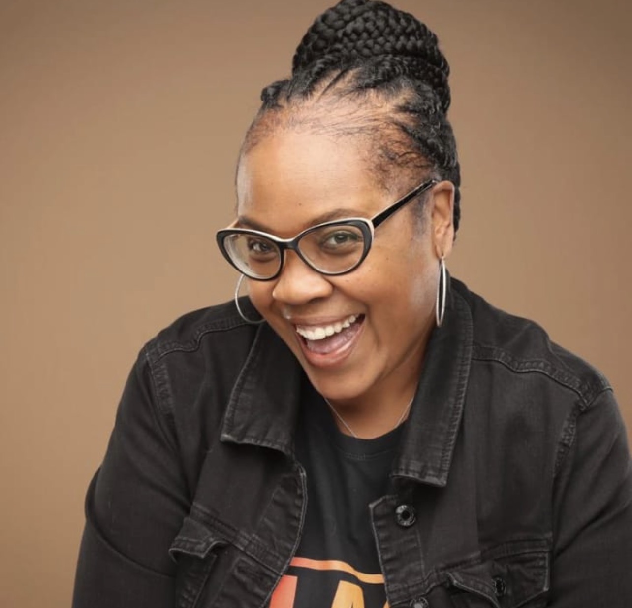 Woman with glasses smiling, wearing a black jacket and hoop earrings, against a plain background.