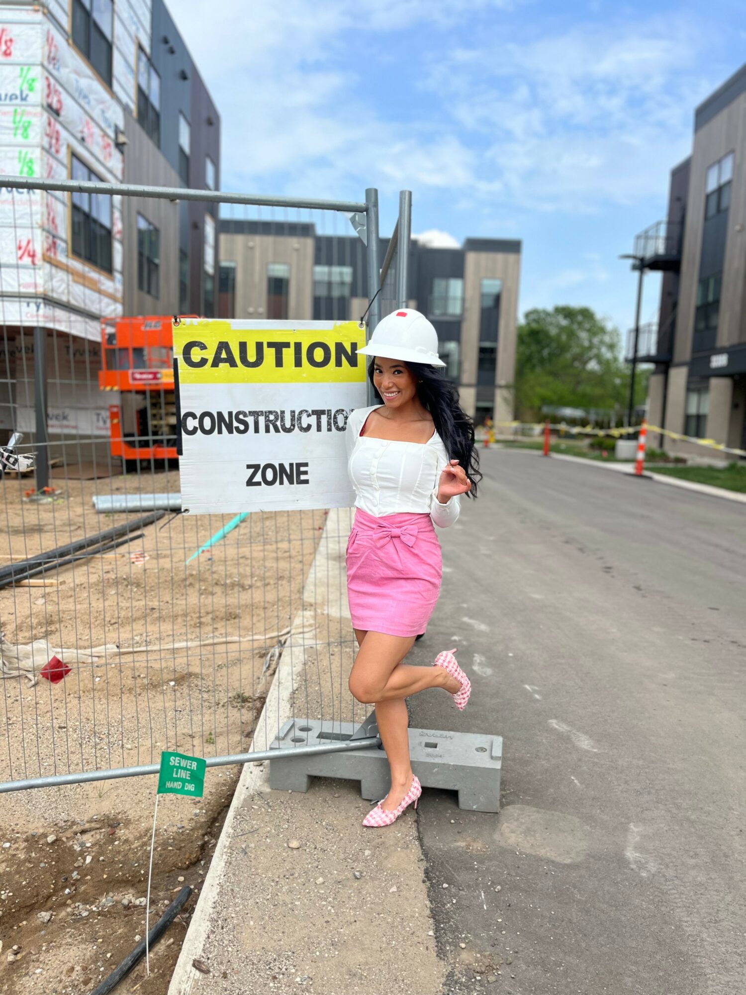Young woman in construction helmet standing near construction site, smiling, on a city street with buildings and blue sky.