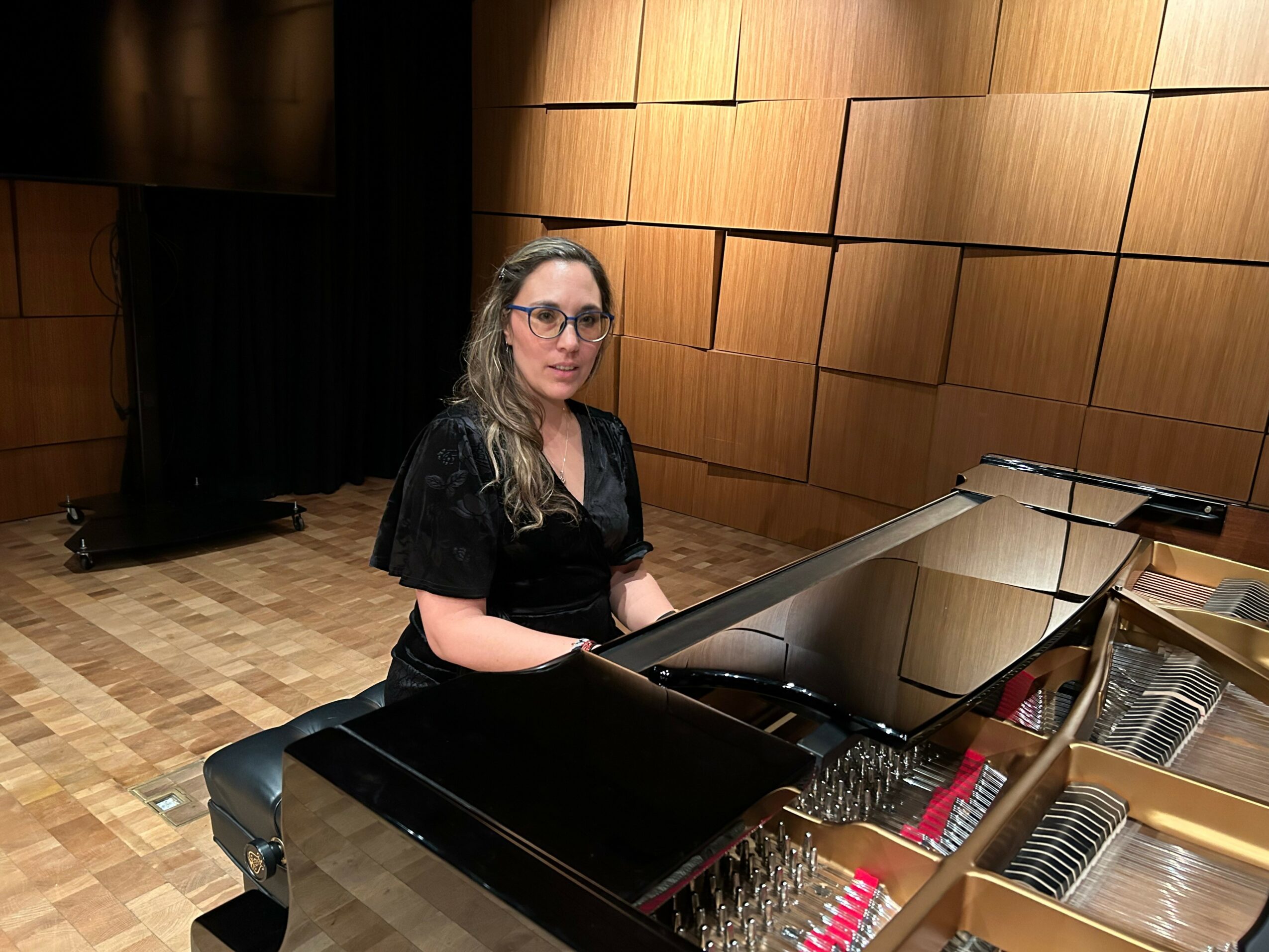 Woman with glasses sitting at a black grand piano in a room with wooden wall panels.