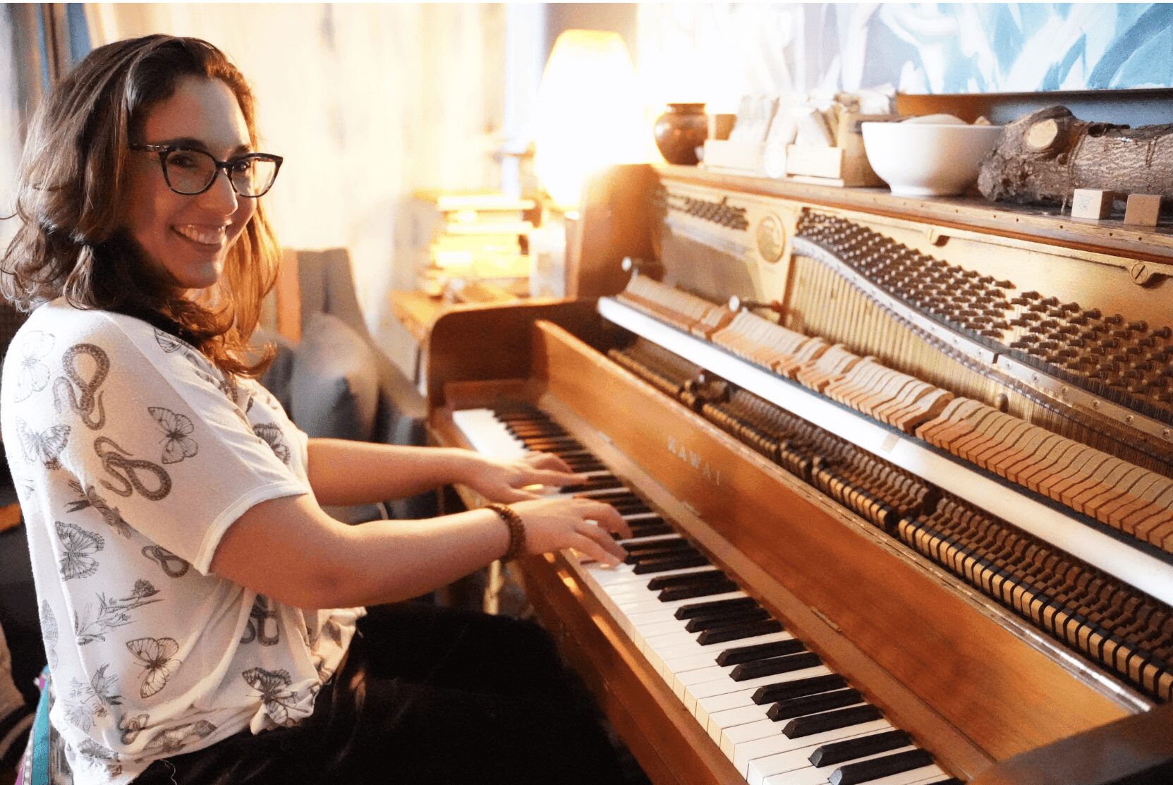Woman playing piano, smiling, in a warmly lit room with a lamp and decorative items, wearing glasses and a patterned shirt.