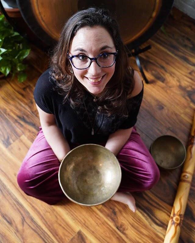 Woman with glasses sitting cross-legged on wooden floor, holding a brass singing bowl, smiling, with plants and furniture nearby.