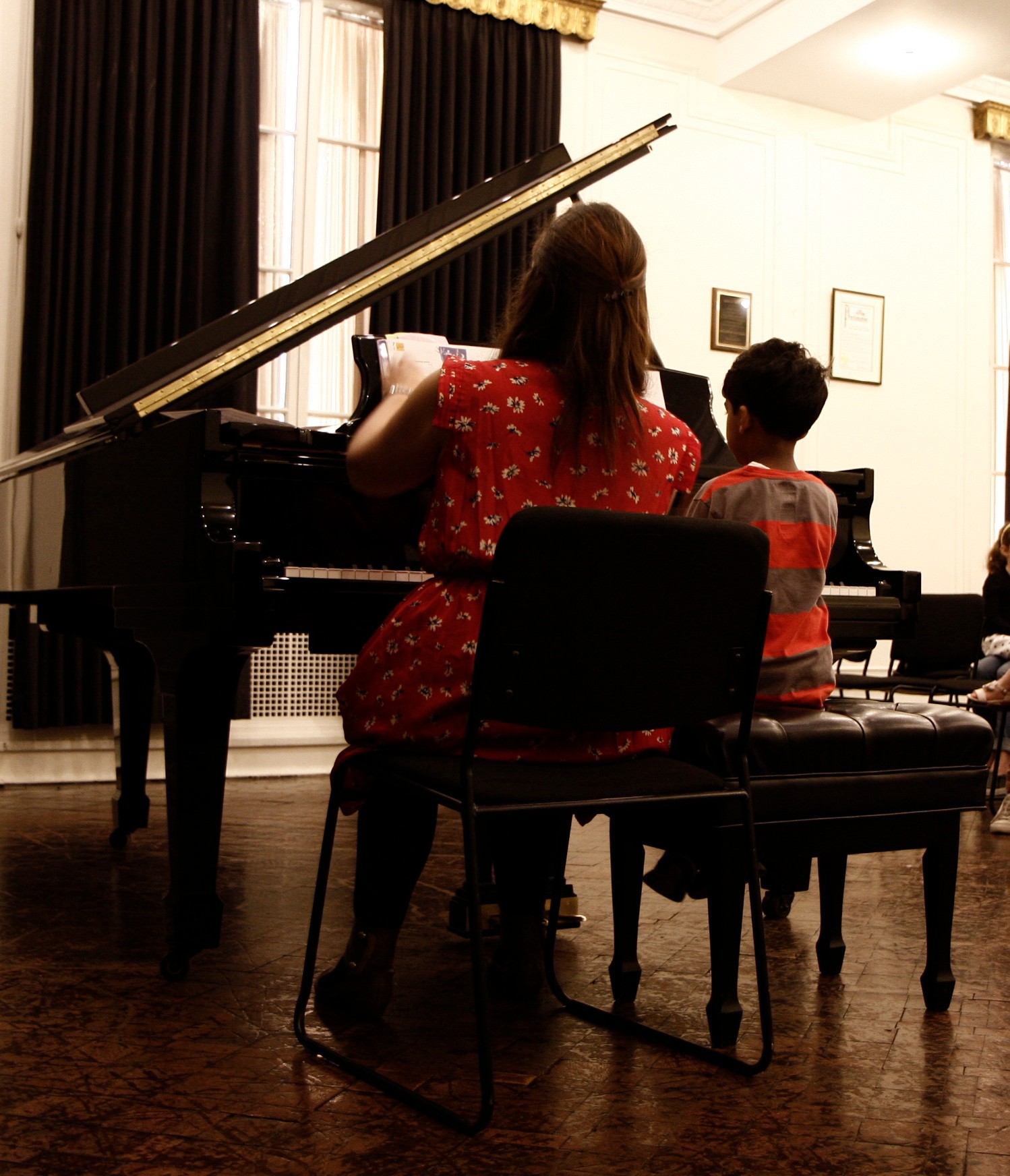 Woman and child sitting at a piano in a room with curtains and framed pictures on the wall.