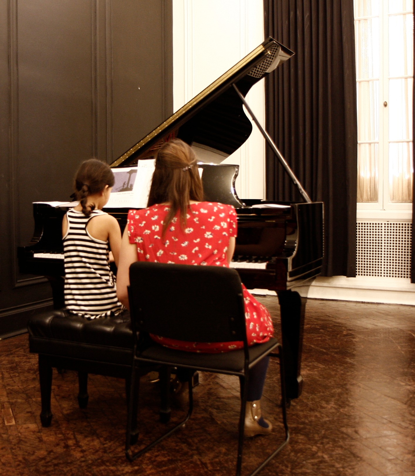 Two children sit at a black grand piano in a room with dark wood floors and curtains, one playing and one watching.