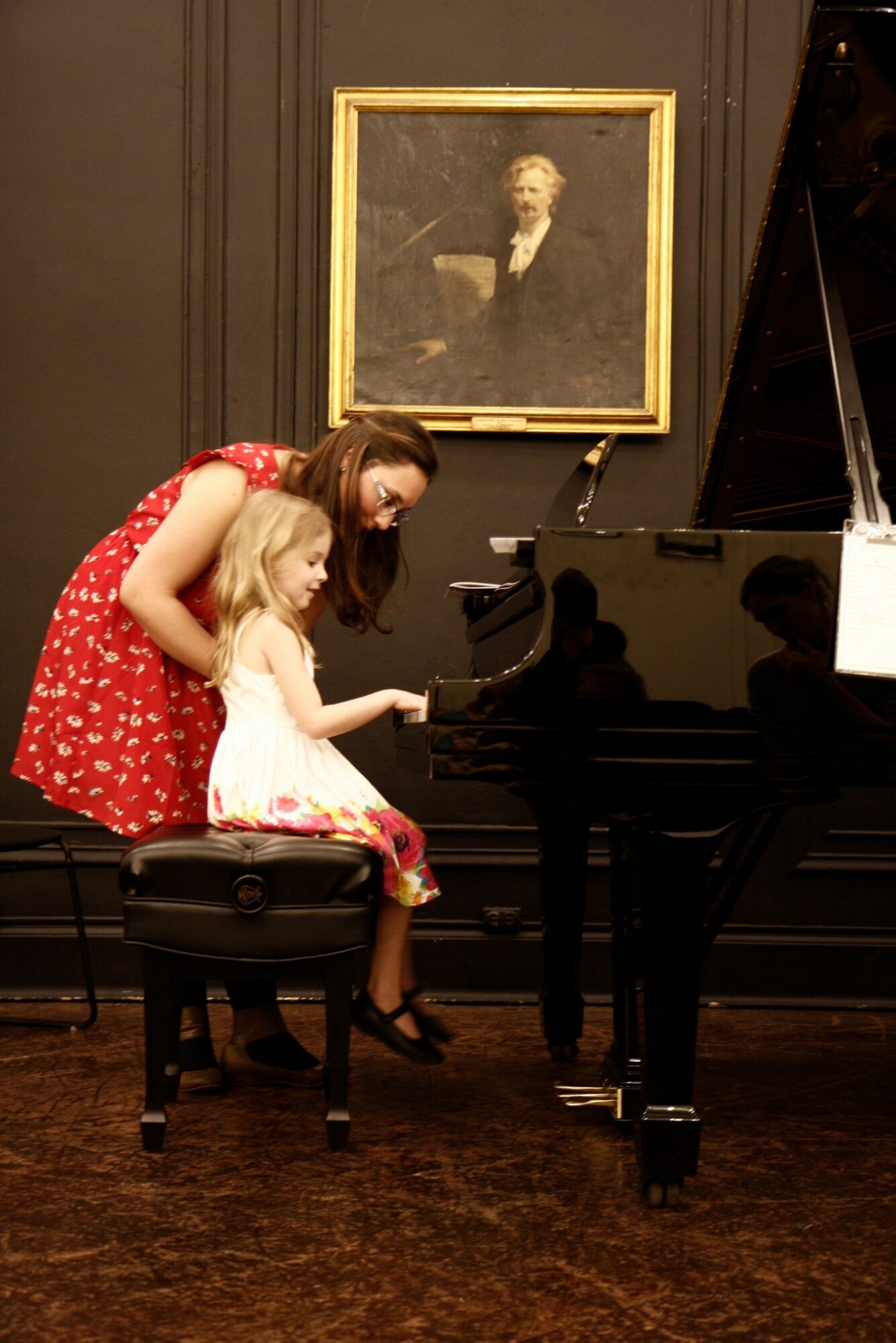 A woman and a young girl sit at a piano, with a portrait hanging on the wall behind them.