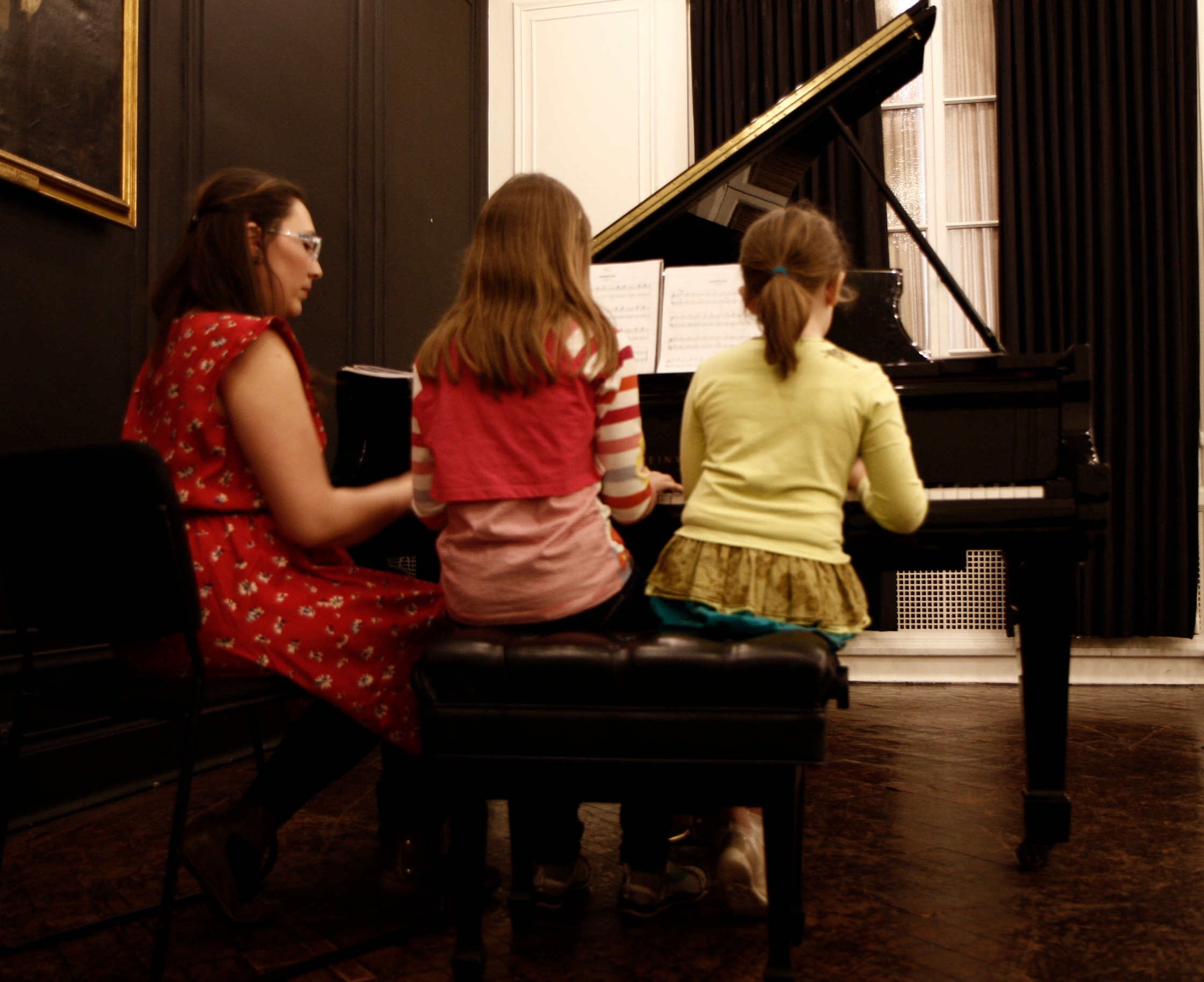 Three girls sit at a grand piano, practicing or playing together in a room with dark walls and wooden floor.